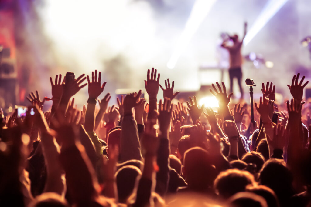 A crowd of people with raised hands at an outdoor concert, with bright stage lights and a performer visible in the background.
