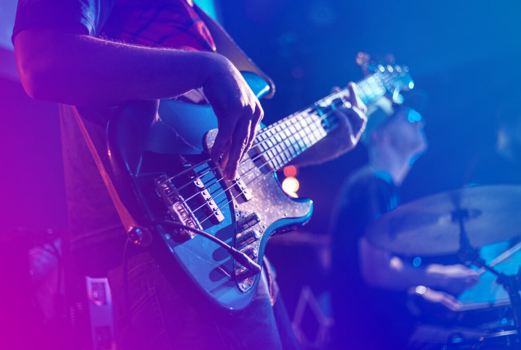 Close-up of a person playing an electric bass guitar on stage, with another musician on drums in the background under colorful lighting.