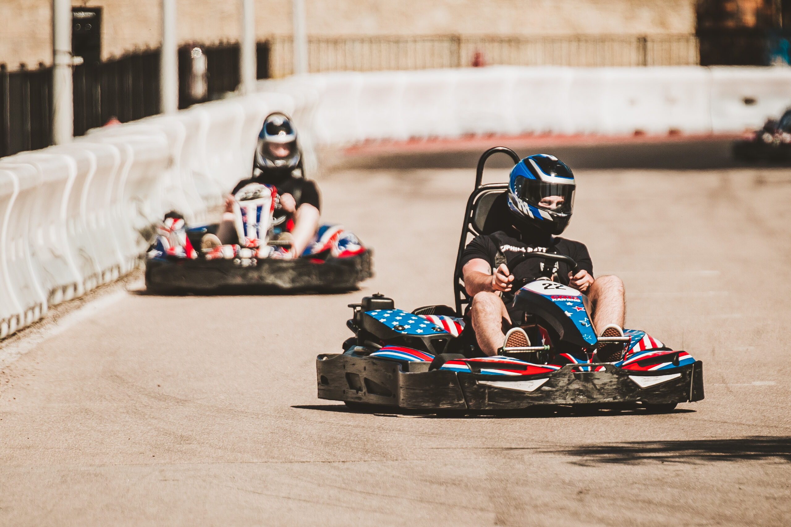Two people wearing helmets drive go-karts on an outdoor track, with one kart ahead of the other. The track is bordered by white barriers.