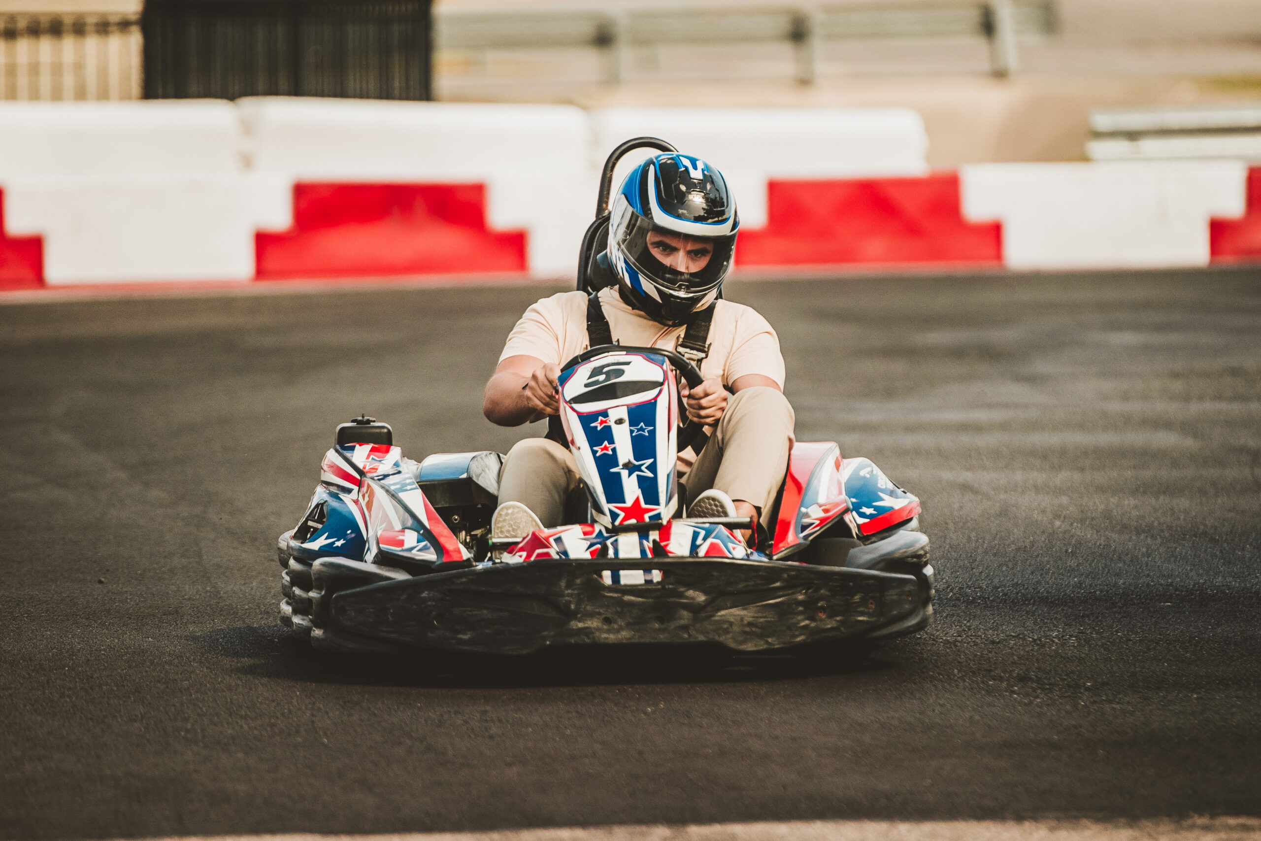 A person wearing a helmet drives a go-kart with a star and stripe design on a paved race track.