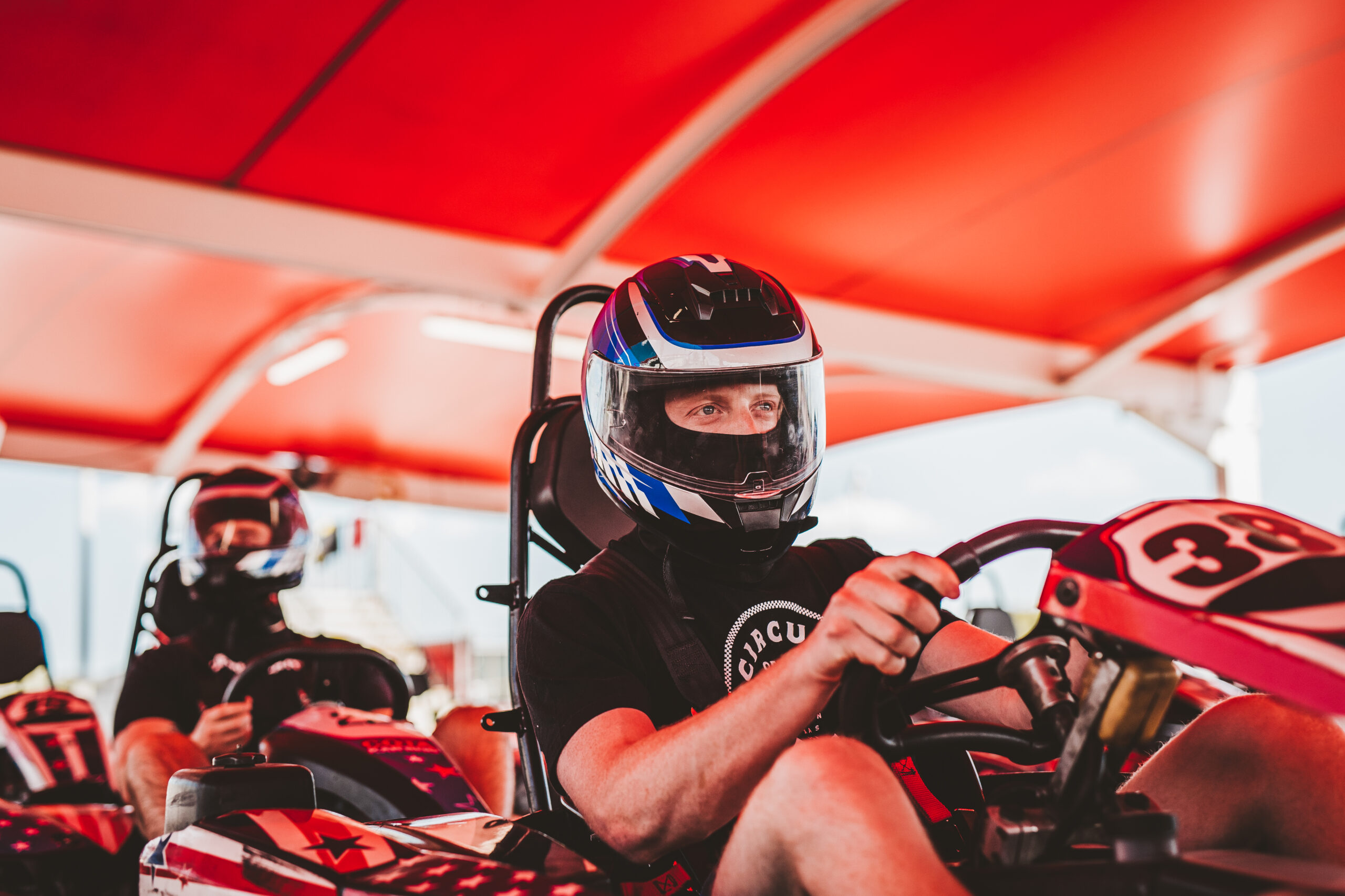 Two people wearing helmets sit in go-karts under a red canopy, with one person in the foreground focused on driving.