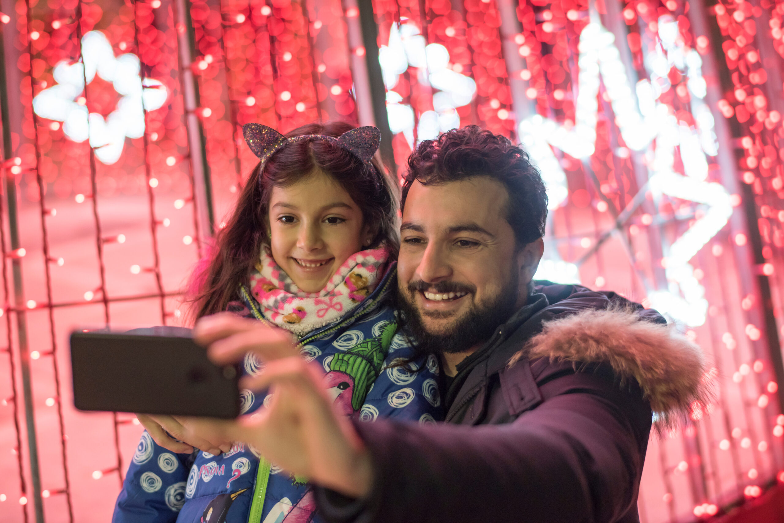 A man and a young girl take a selfie together in front of a backdrop of red lights and illuminated star shapes.