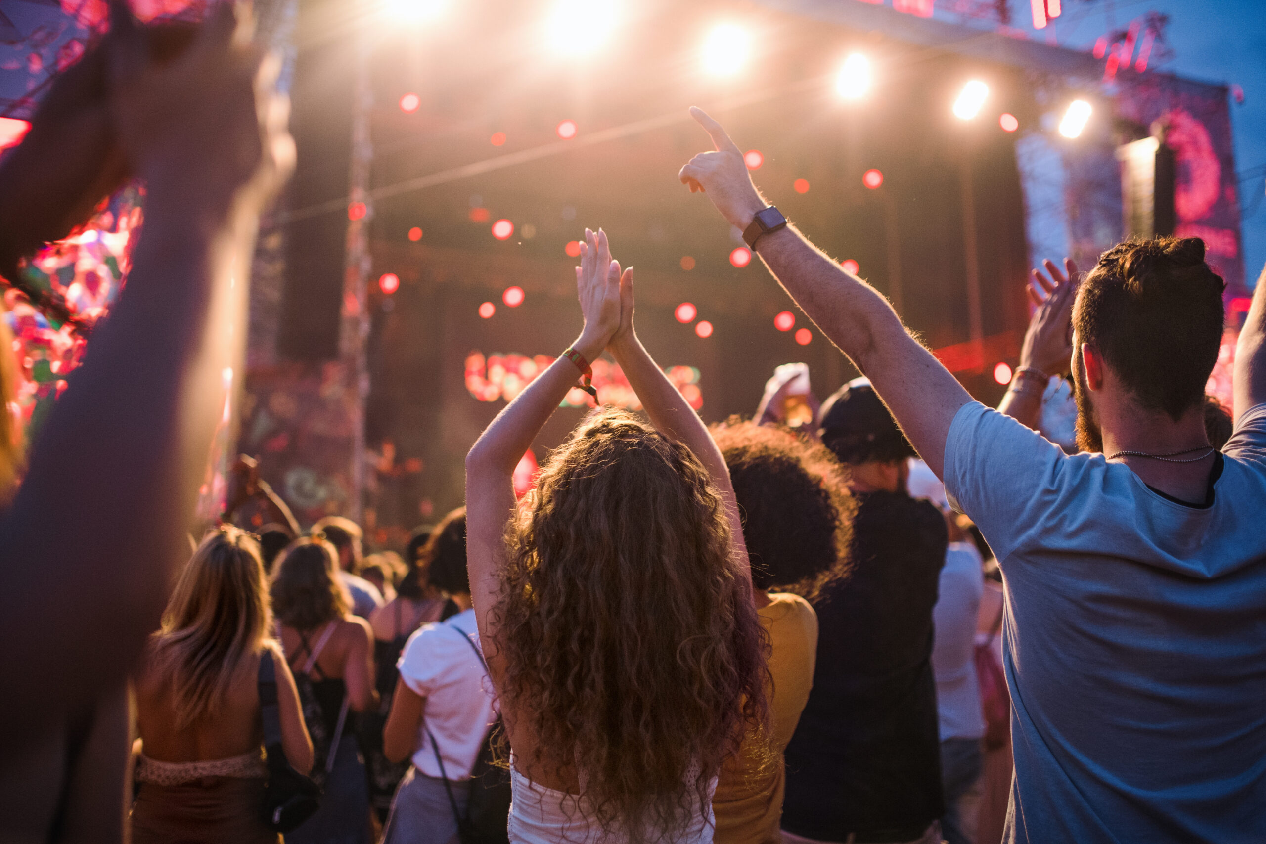 A crowd of people with raised arms face a brightly lit outdoor concert stage during a live music performance.