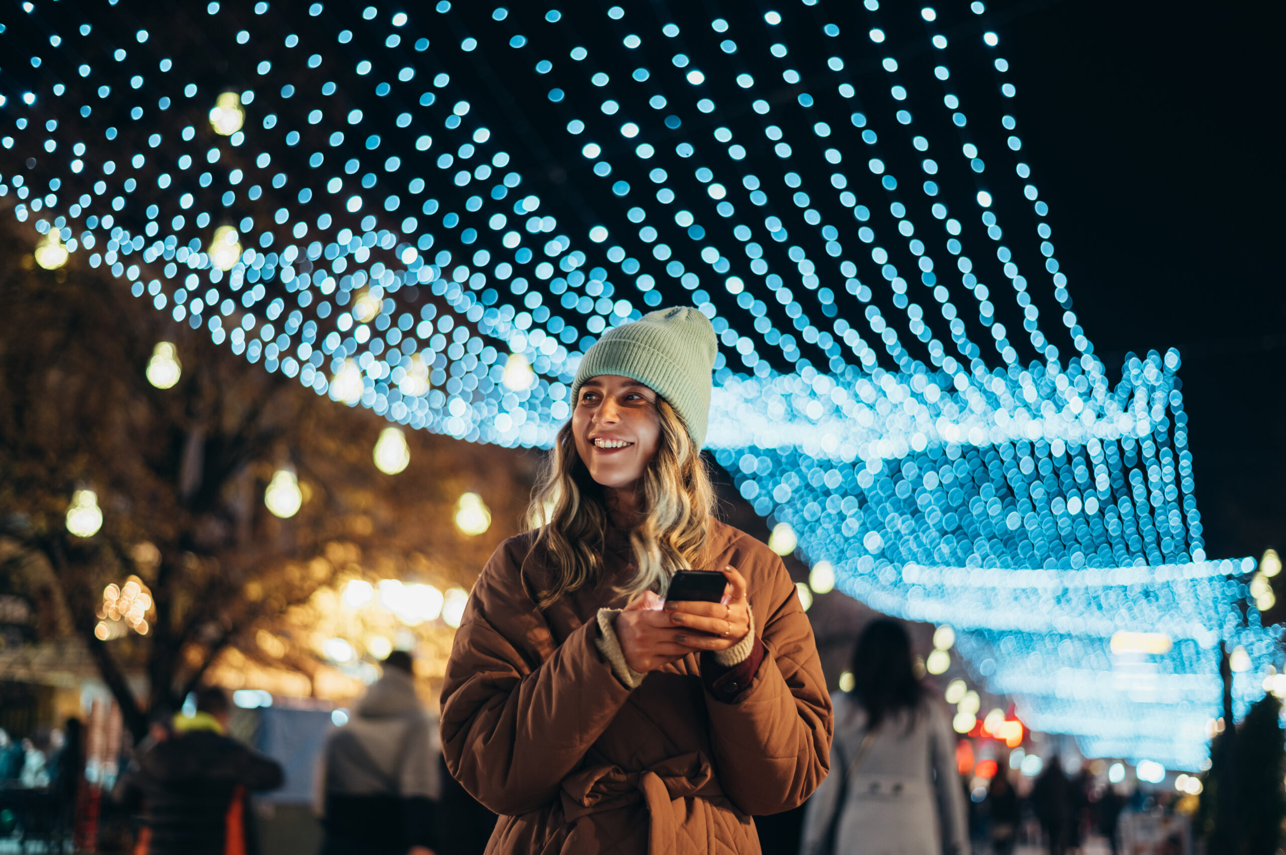 A woman in a winter coat and beanie holds a smartphone and smiles, standing under blue string lights at night with blurred people in the background.