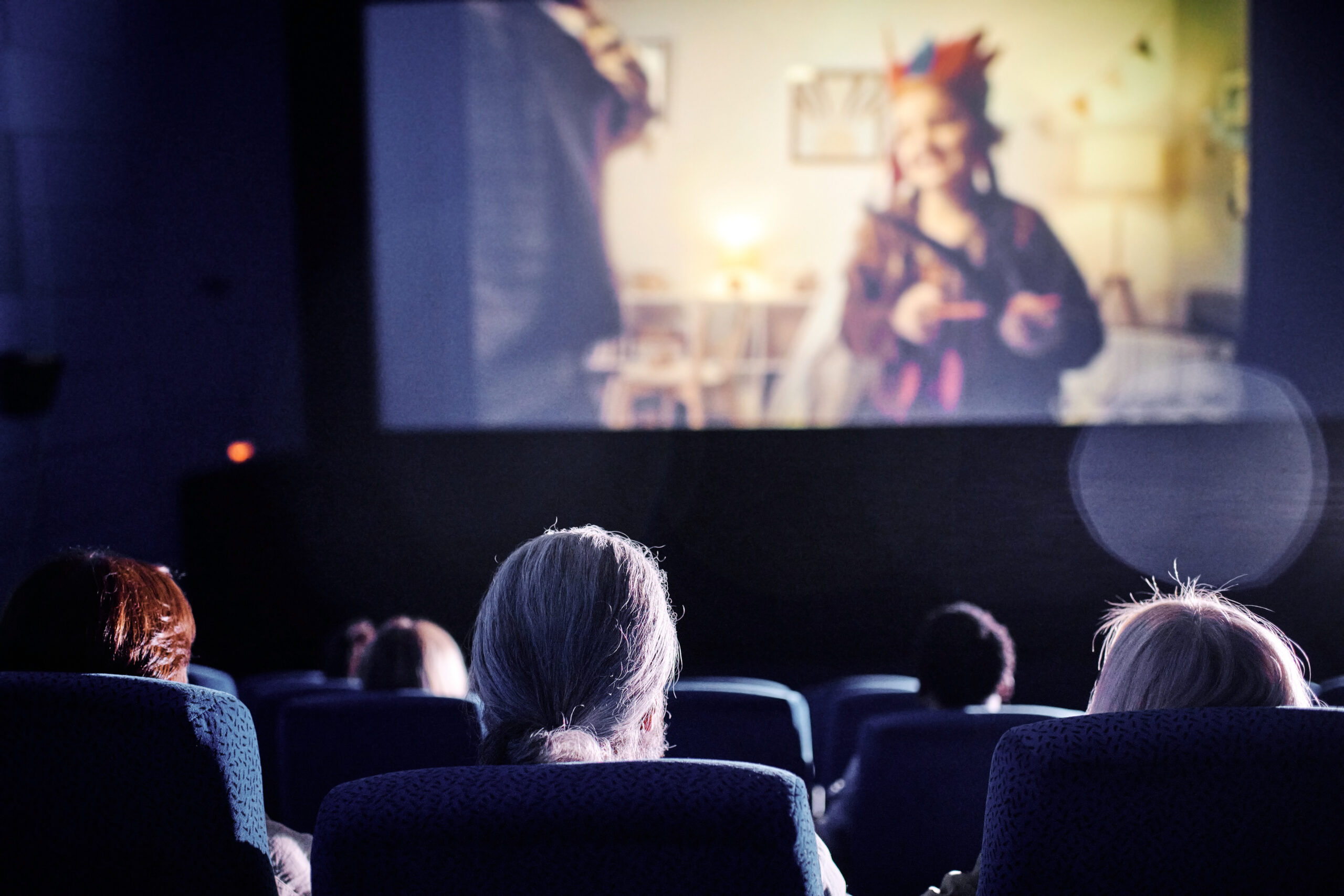People sitting in a dark movie theater facing a screen showing a blurry image of a person in colorful attire.