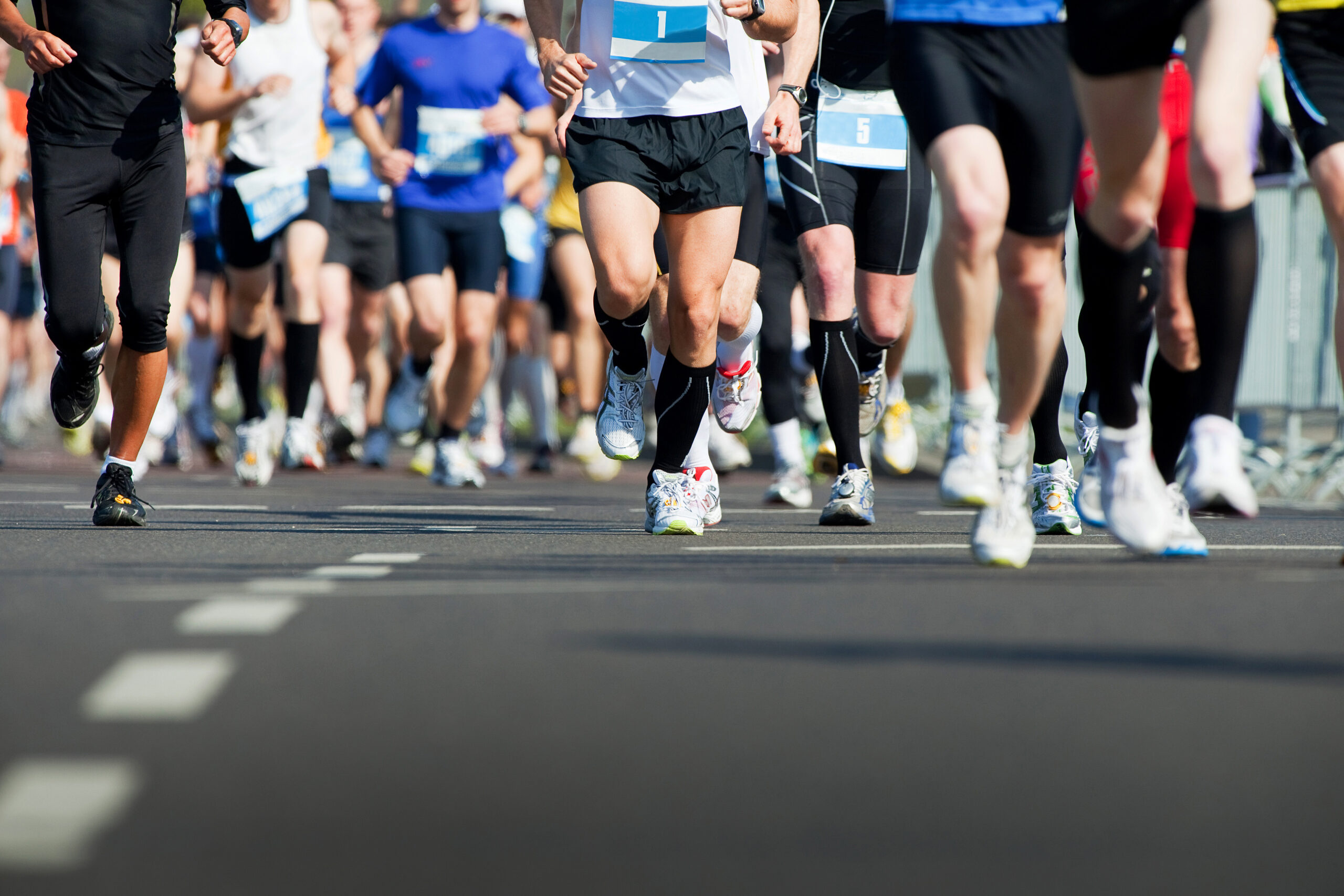 A group of runners in athletic gear participate in a road race, with only their legs and lower bodies visible as they run on a paved street.