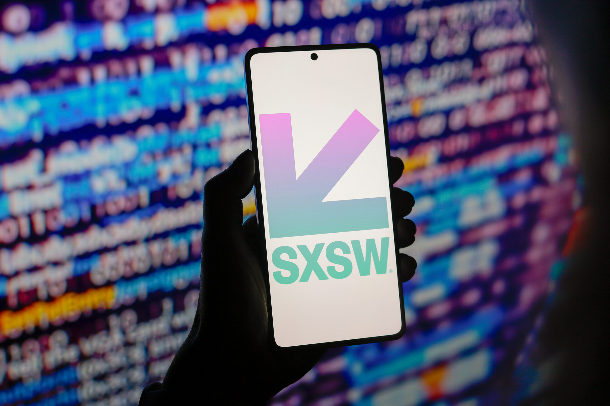 A hand holds a smartphone displaying the SXSW logo, with a screen of blurred multicolored code in the background.