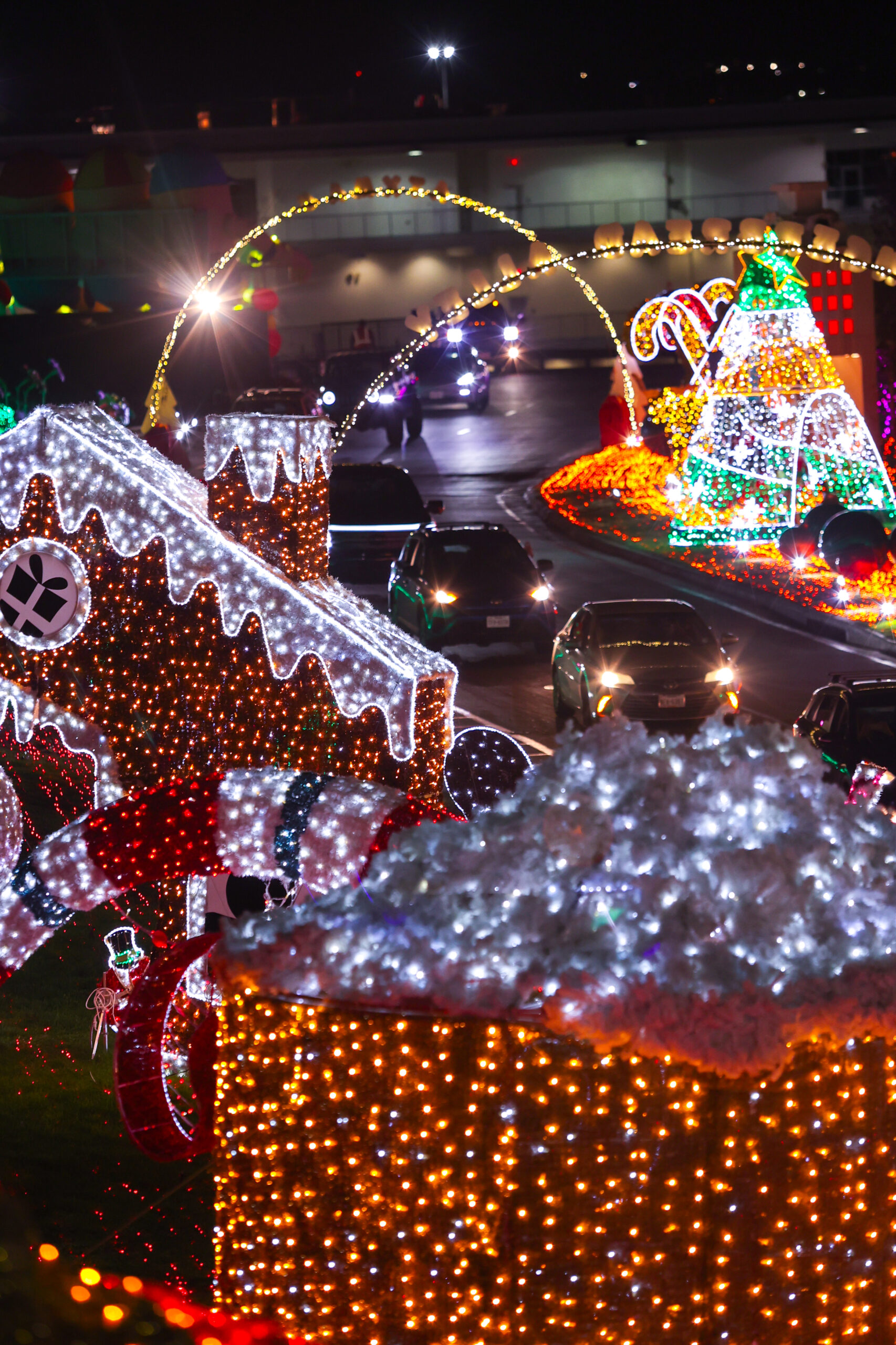 Cars drive through a holiday light display featuring gingerbread houses, candy canes, and a large Christmas tree illuminated with colorful lights at night.