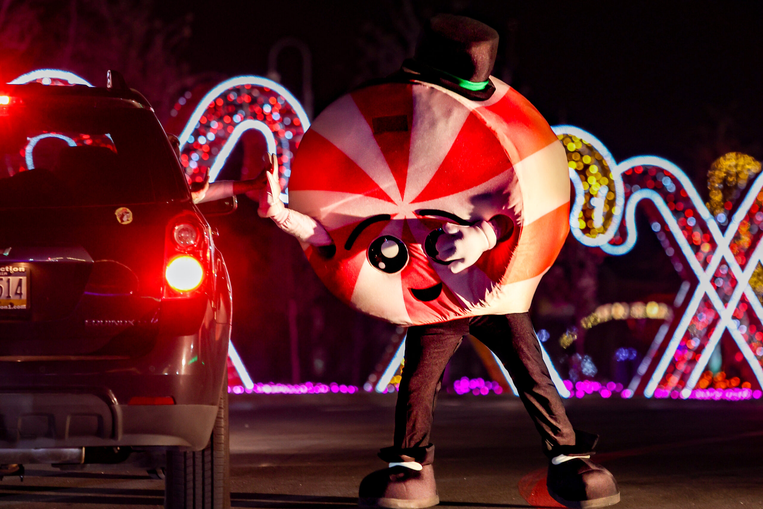 A person in a peppermint candy costume waves at a car during a nighttime holiday light display with candy cane decorations in the background.