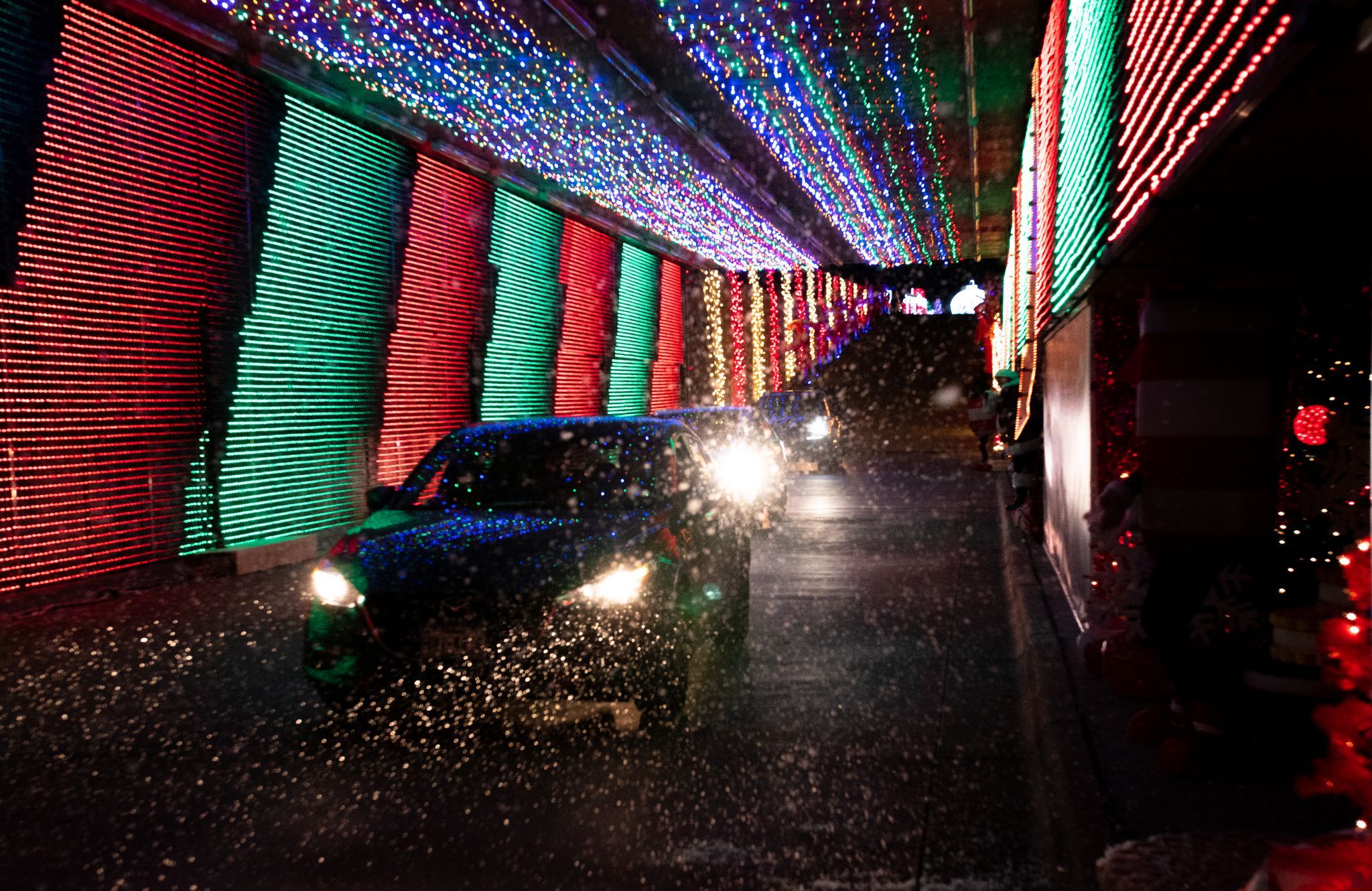 Cars drive through a tunnel decorated with rows of red, green, and blue holiday lights, seen through a rain-splattered window at night.