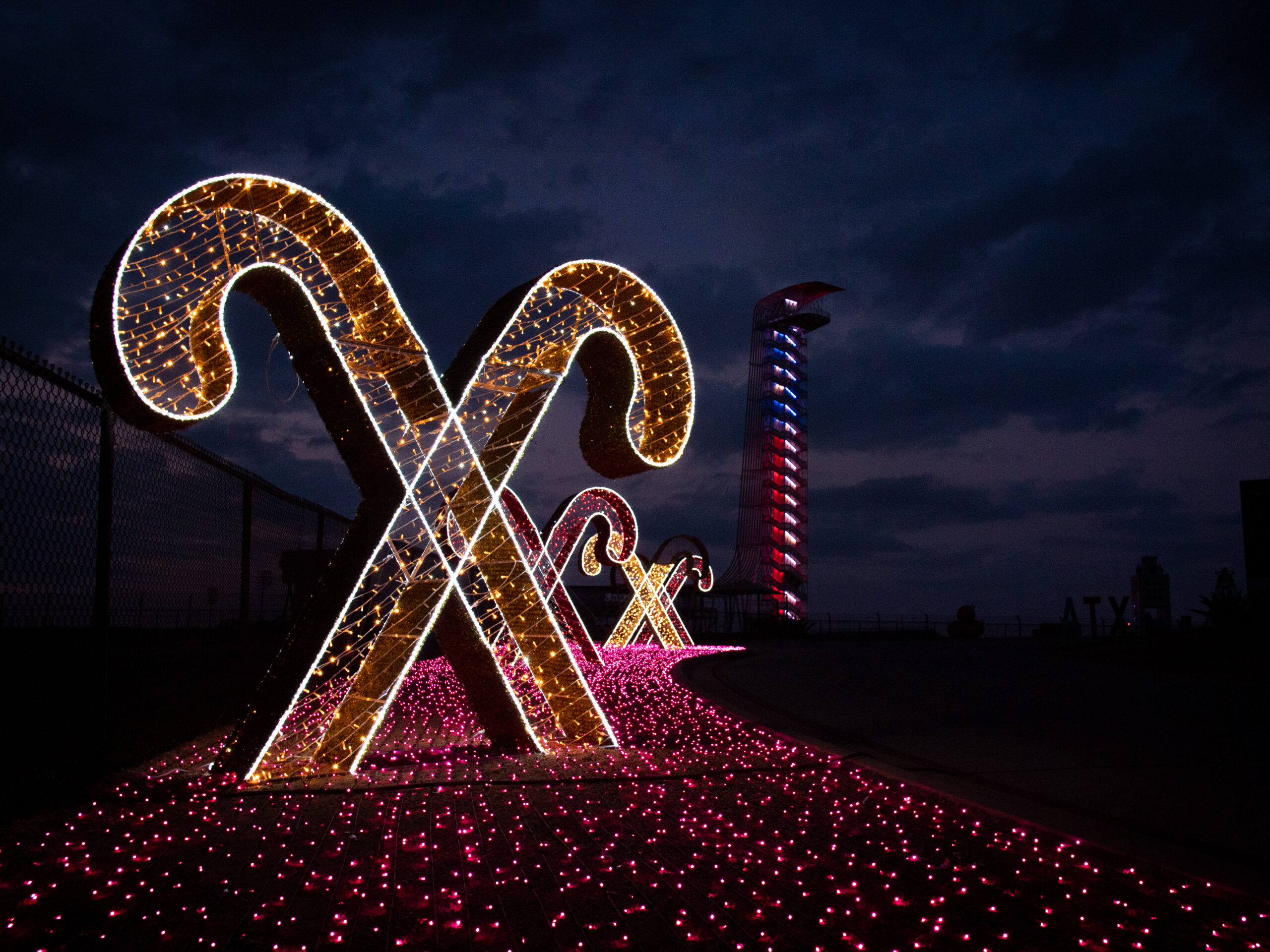 Large illuminated candy cane decorations line a path with pink lights at night, with a tall tower lit in red, white, and blue in the background.