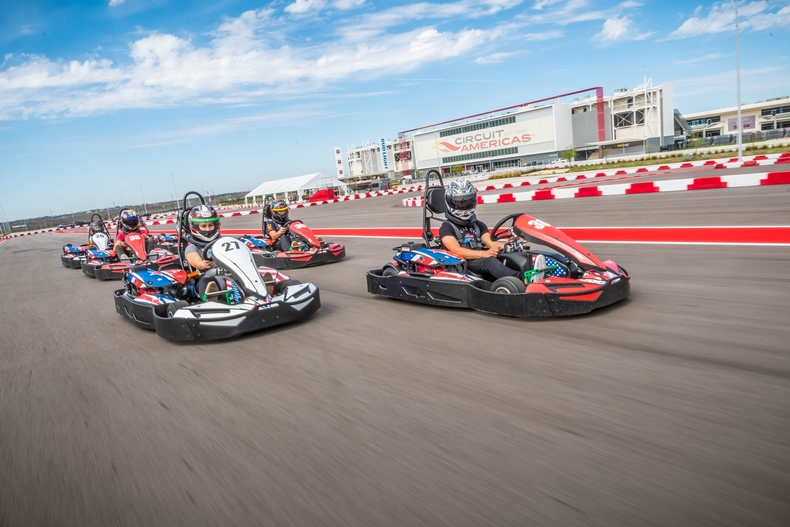 Four people race go-karts on an outdoor track with the Circuit of the Americas building visible in the background under a blue sky.