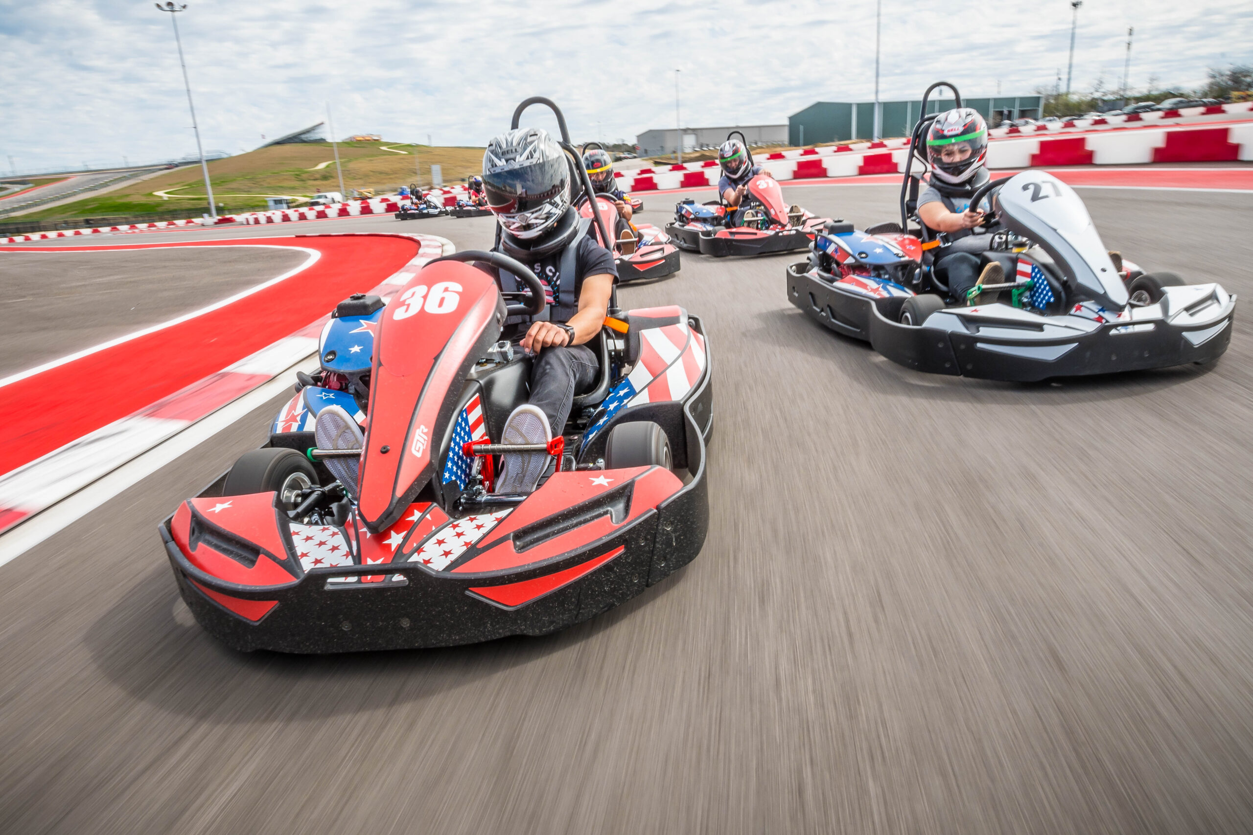 Several people wearing helmets race go-karts on an outdoor track with red and white barriers. The lead kart is number 36.