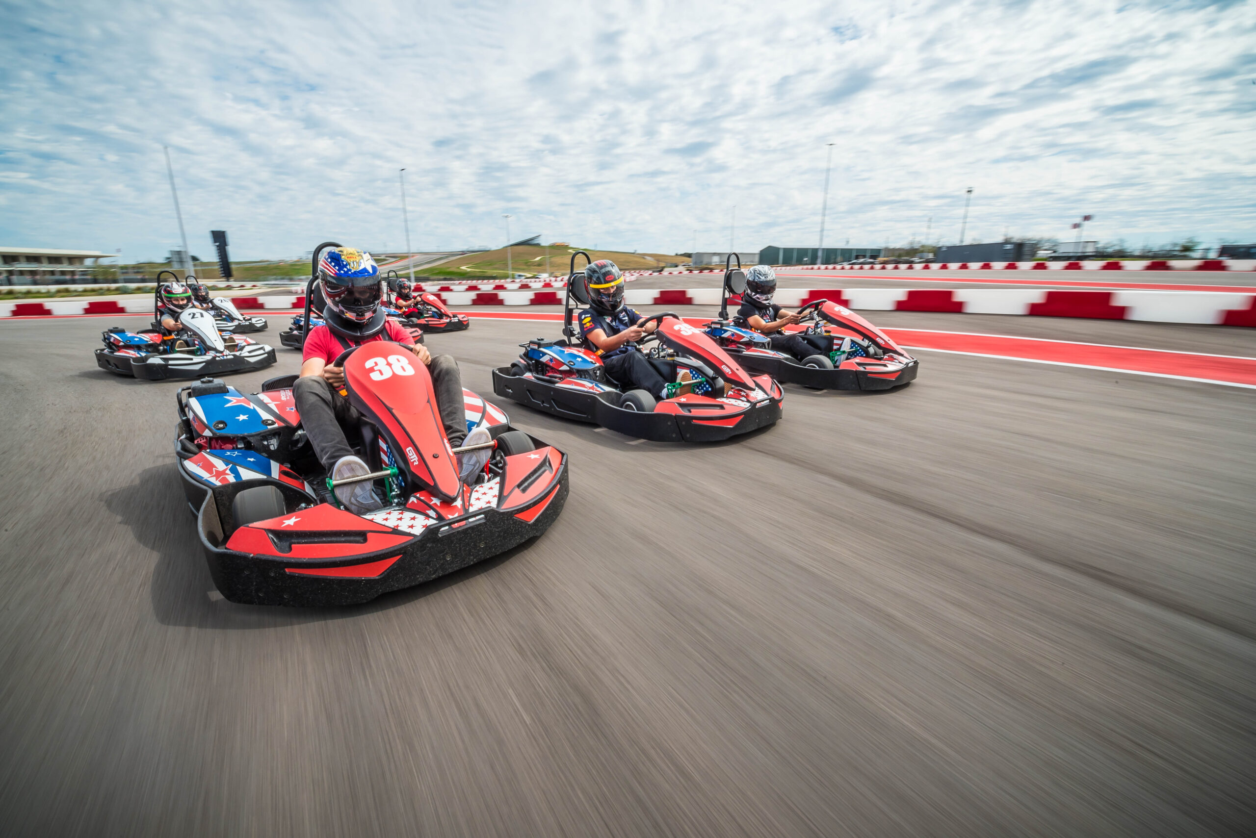 Four people in helmets race red and black go-karts on an outdoor track under a partly cloudy sky. The driver in kart number 38 is in the lead.