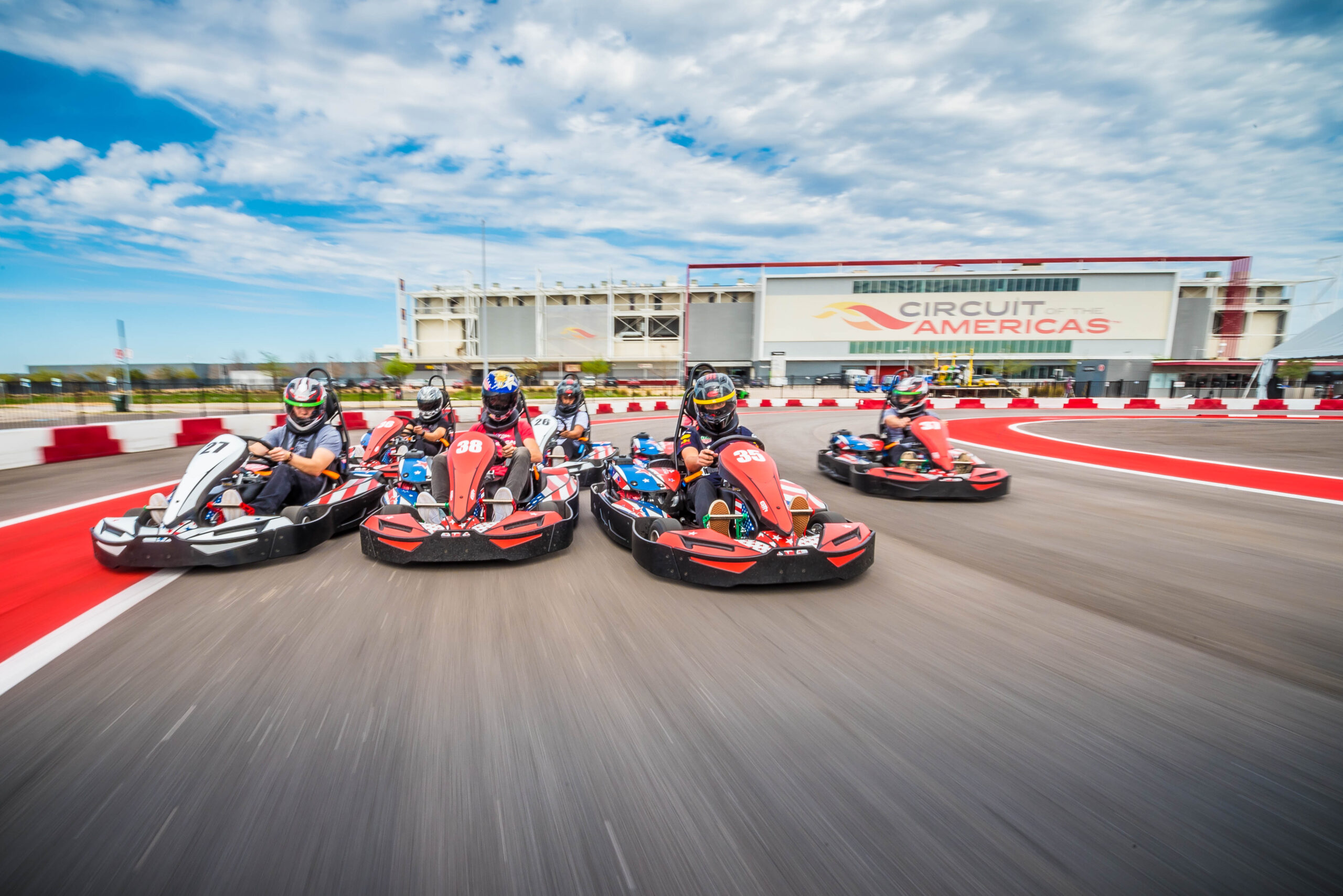 Four people in go-karts race on an outdoor track in front of a building labeled "Circuit of the Americas" under a partly cloudy sky.
