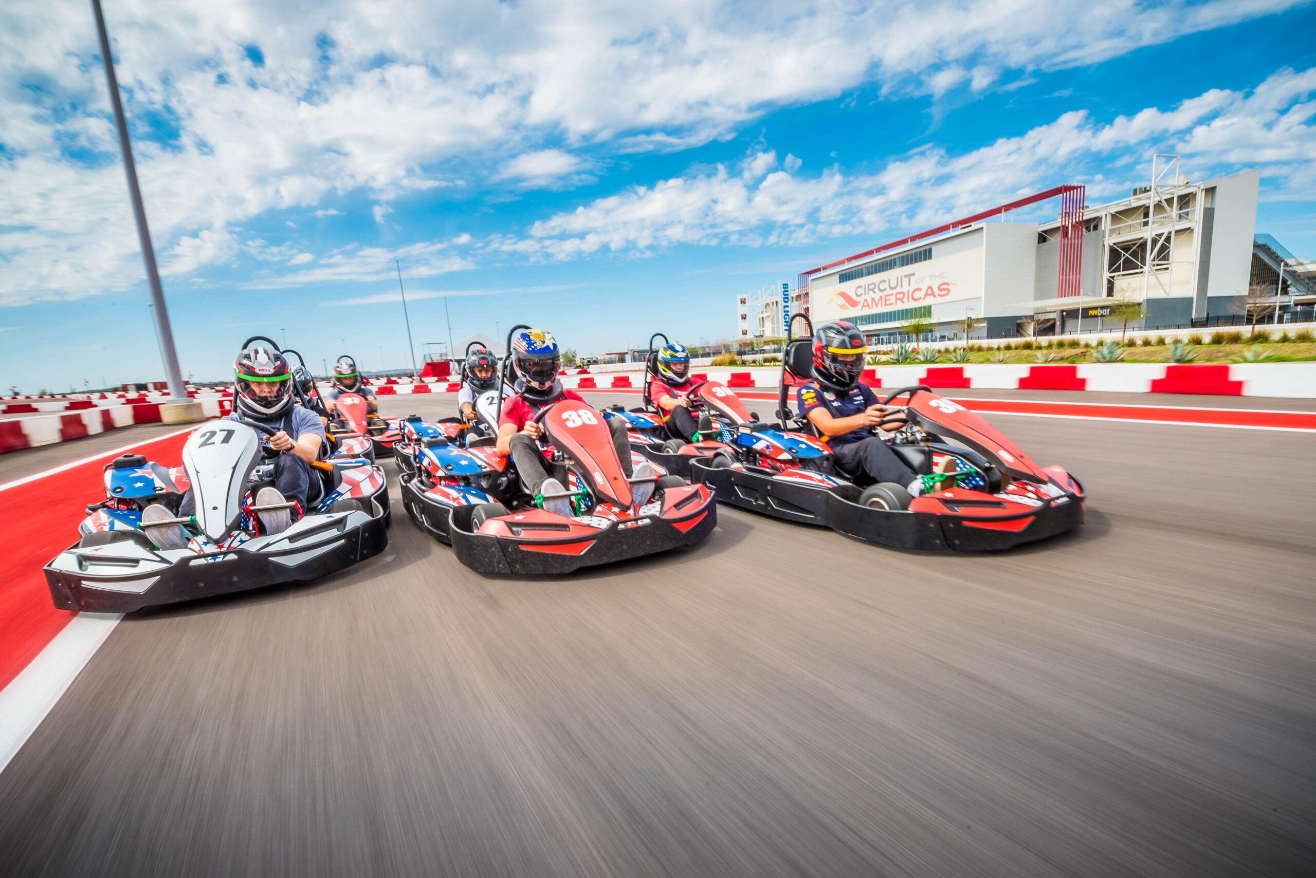 A group of people racing go-karts on an outdoor track with the Circuit of the Americas building visible in the background under a partly cloudy sky.