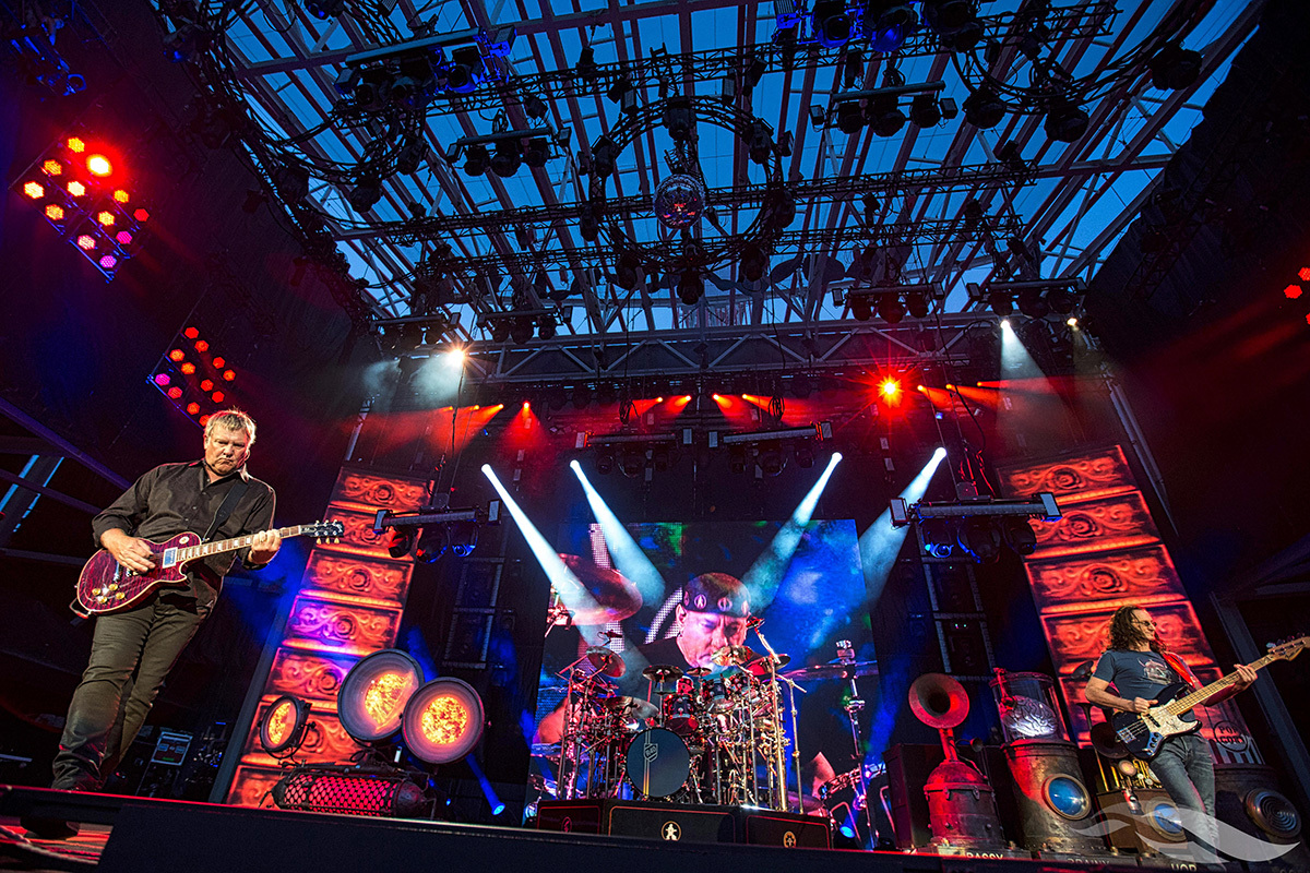 Three musicians perform on a brightly lit stage, with a drummer centered in front of a large screen displaying his image and guitarists on either side playing electric instruments.