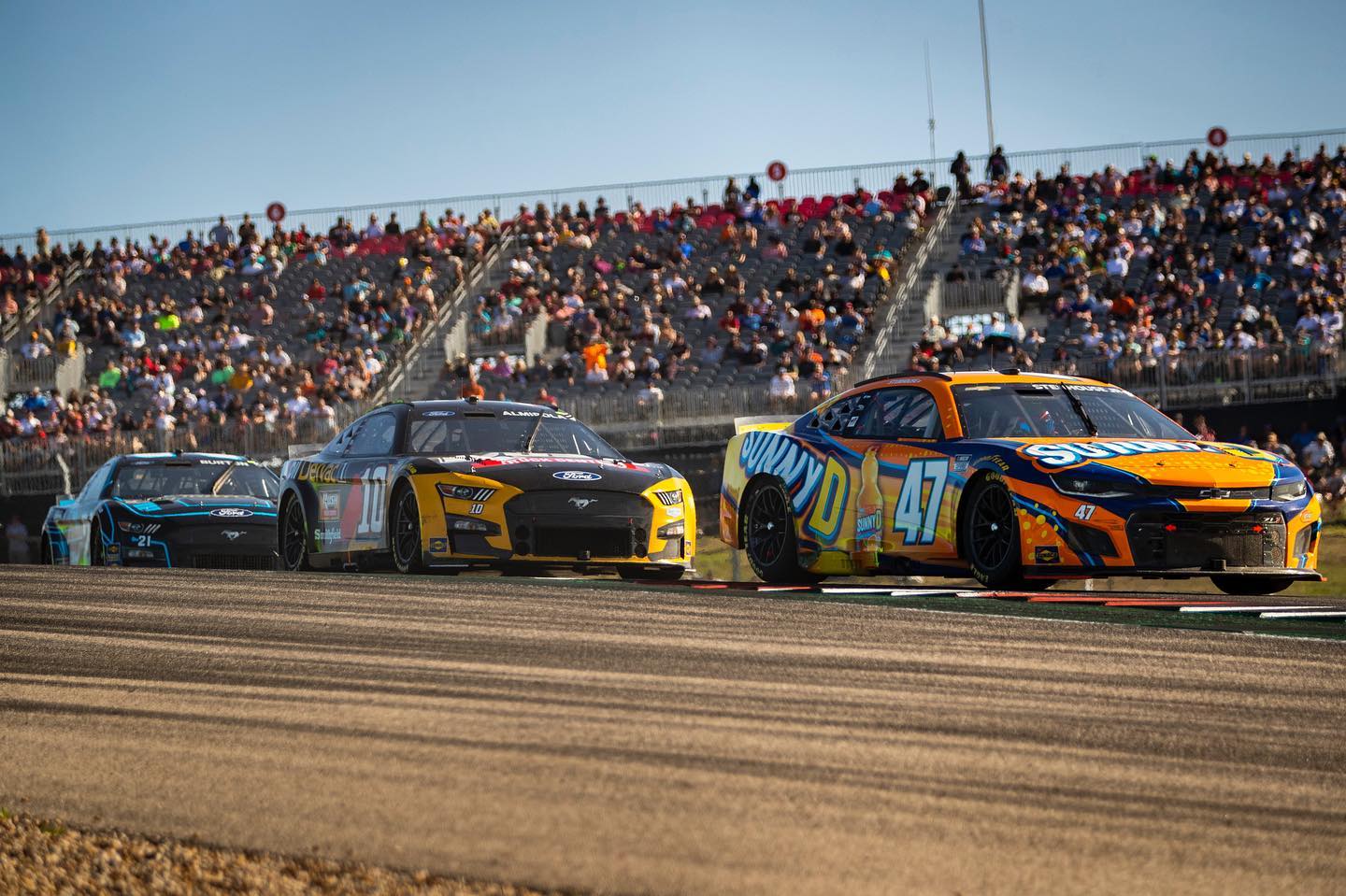 Three stock cars race on a track in front of a packed grandstand during a motorsports event on a sunny day.