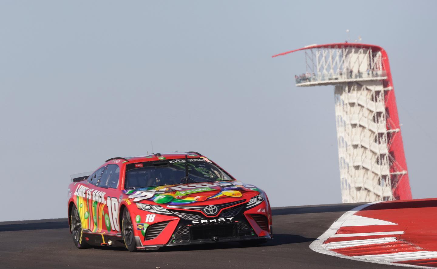 A red NASCAR race car with colorful candy graphics on the hood drives on a racetrack with a tall observation tower in the background.