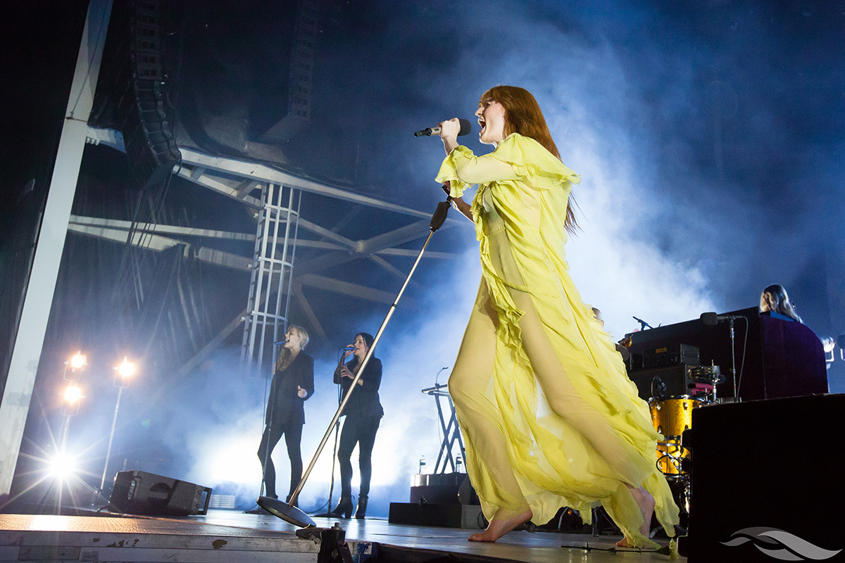 A barefoot singer in a flowing yellow dress performs energetically on stage, with band members and musical equipment in the background and dramatic lighting.