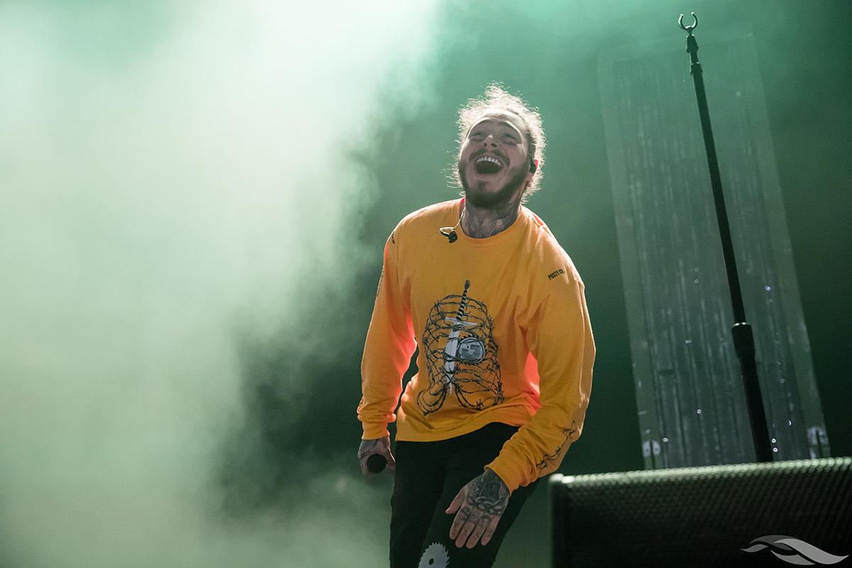 A performer in a yellow long-sleeve shirt holding a microphone sings on stage amid smoke and green lighting.