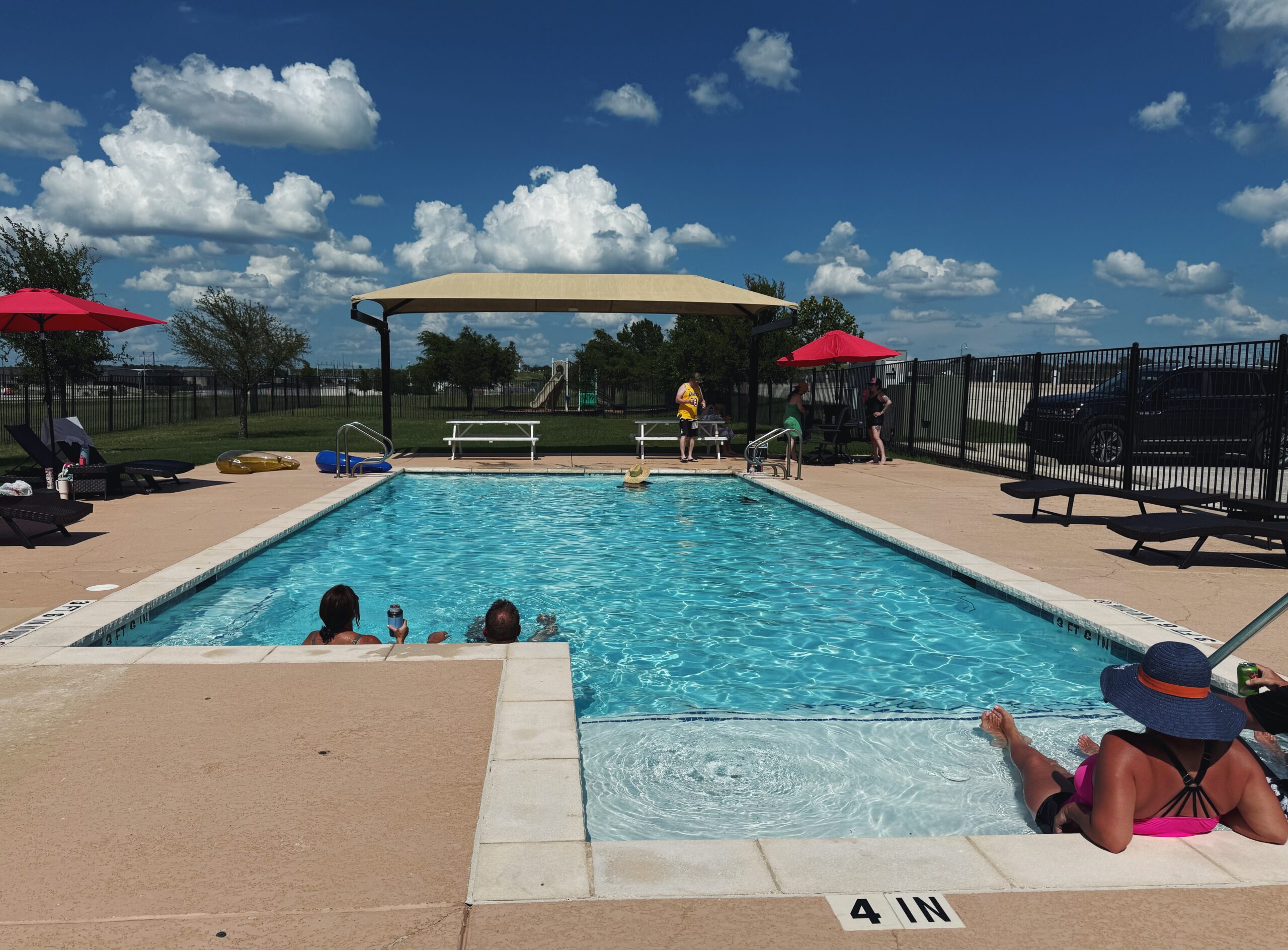 Outdoor pool on a sunny day with several people relaxing in and around the water; lounge chairs, umbrellas, and a canopy are visible.