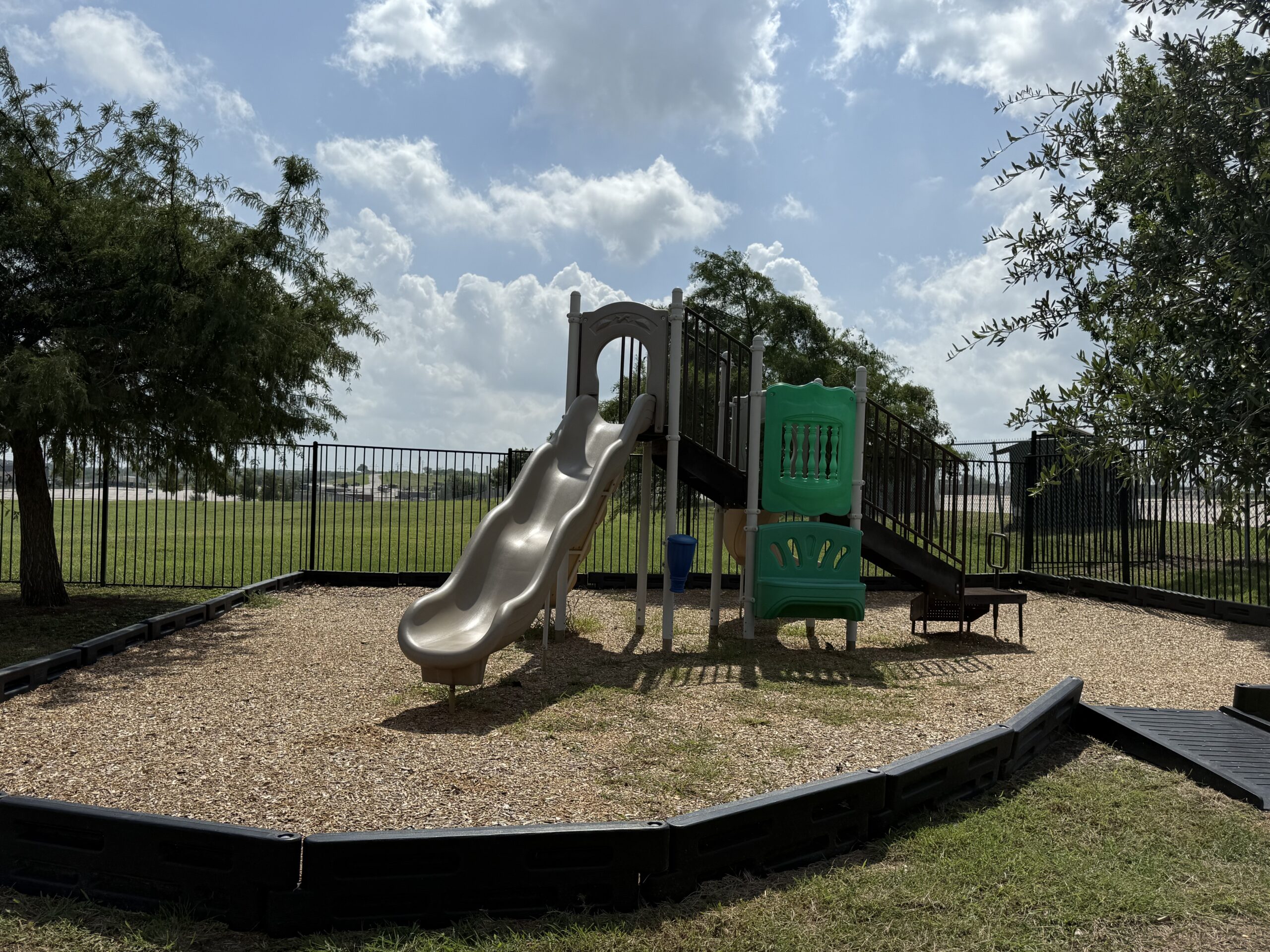 A playground with a slide, ladder, and green climbing structure set on wood chips, enclosed by a black metal fence, under a partly cloudy sky.