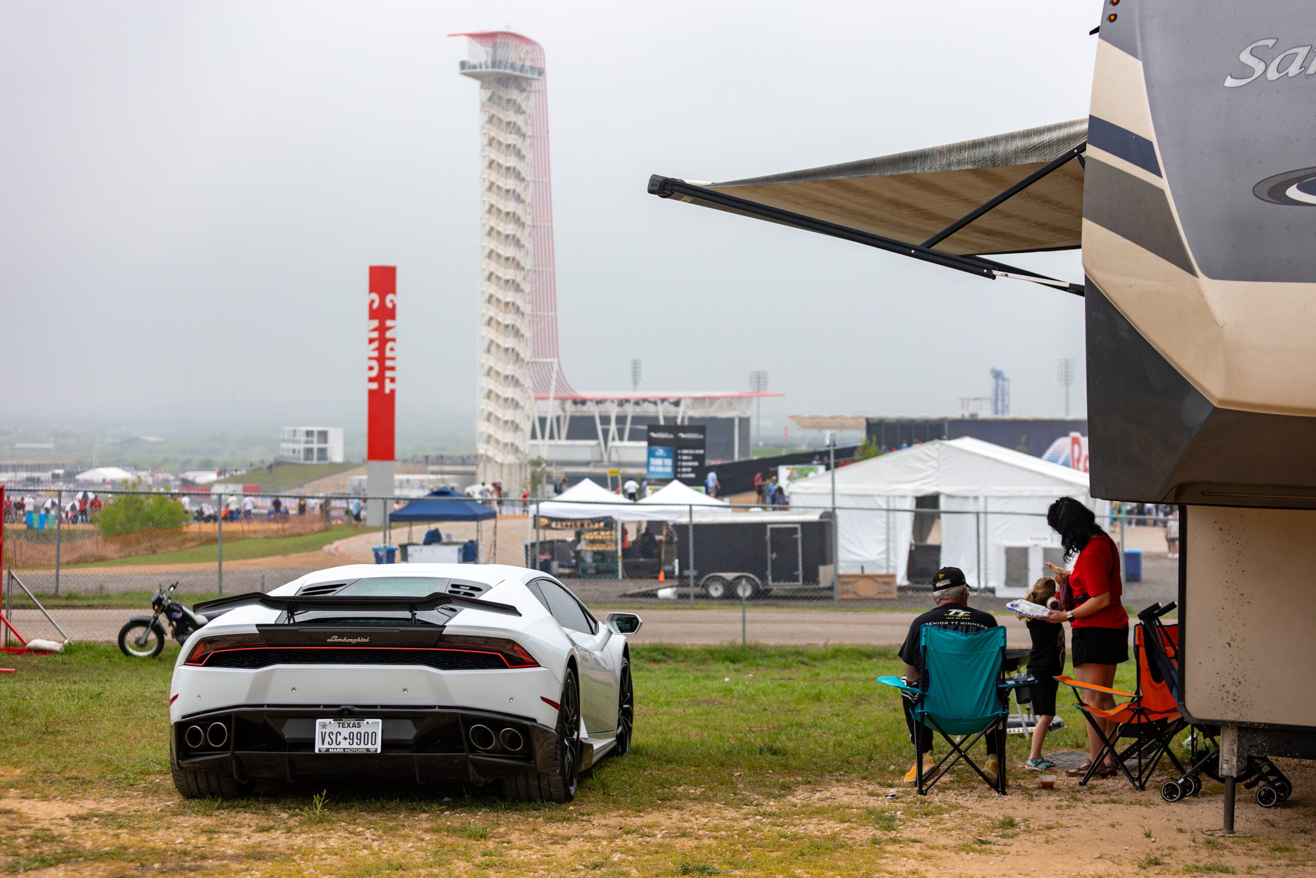 A white Lamborghini is parked on grass near an RV, with three people sitting beside it. In the background, the Circuit of the Americas tower and event tents are visible.