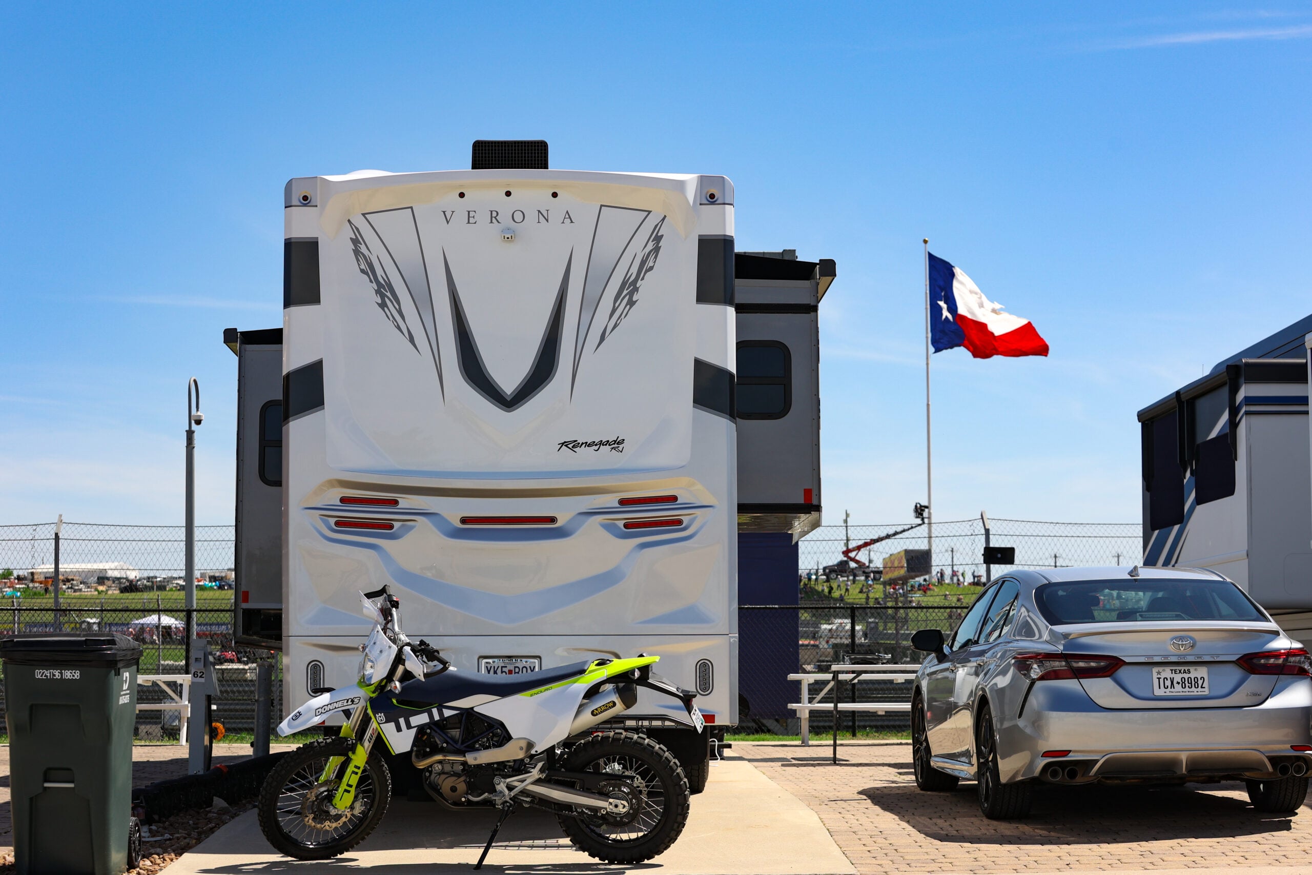 A dirt bike and a car are parked behind a large RV at a campground, with a Texas state flag visible in the background.