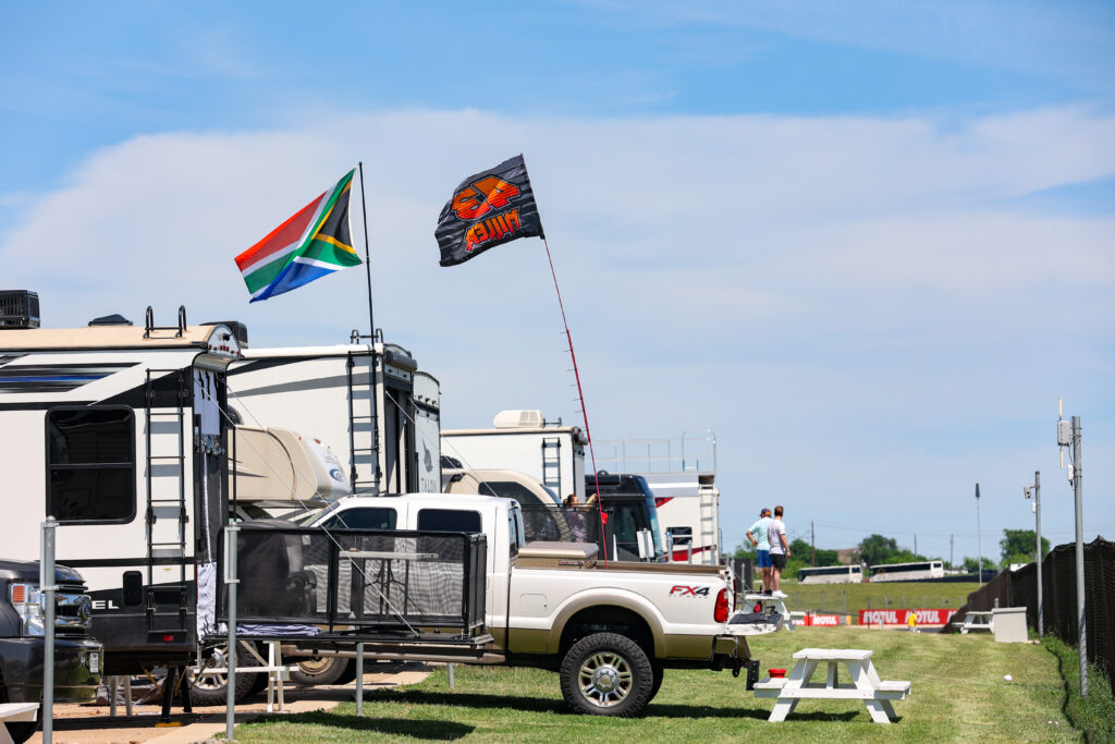 Several RVs and a pickup truck are parked on grass. A South African flag and another black flag are flying. Two people stand in the background near a fence.
