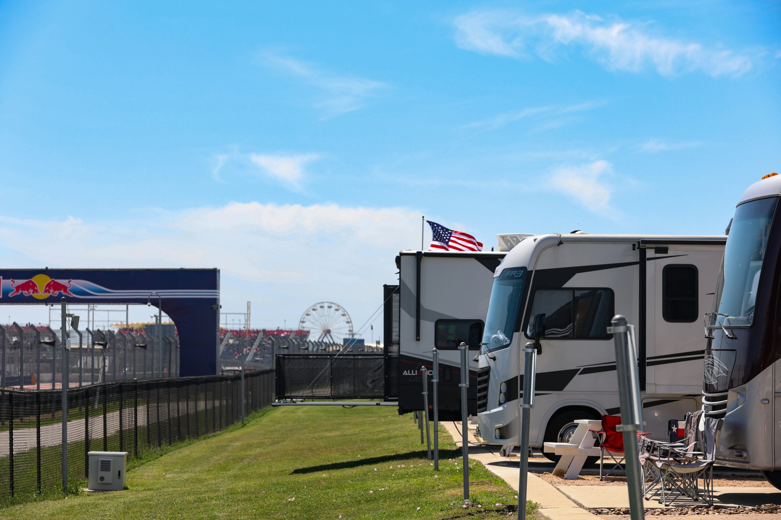 RVs are parked along a fence near a racetrack, with an American flag and a ferris wheel visible in the background under a clear blue sky.