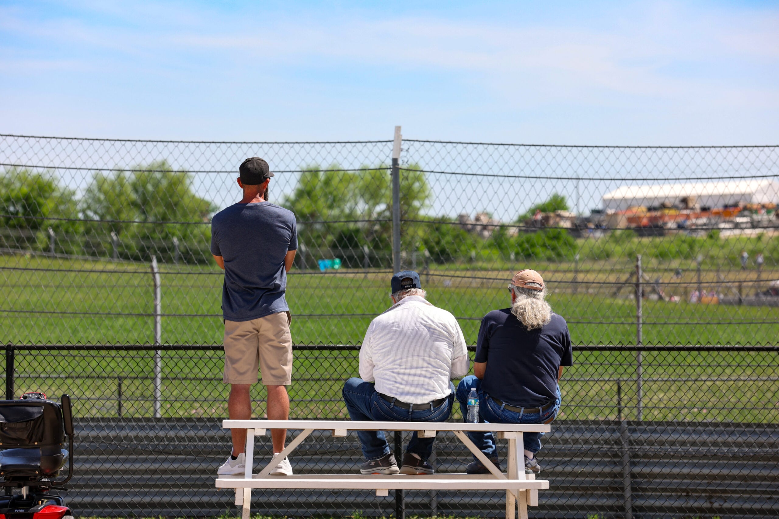 Three people watch a racetrack from bleachers behind a chain-link fence on a sunny day; one is standing while two are seated.