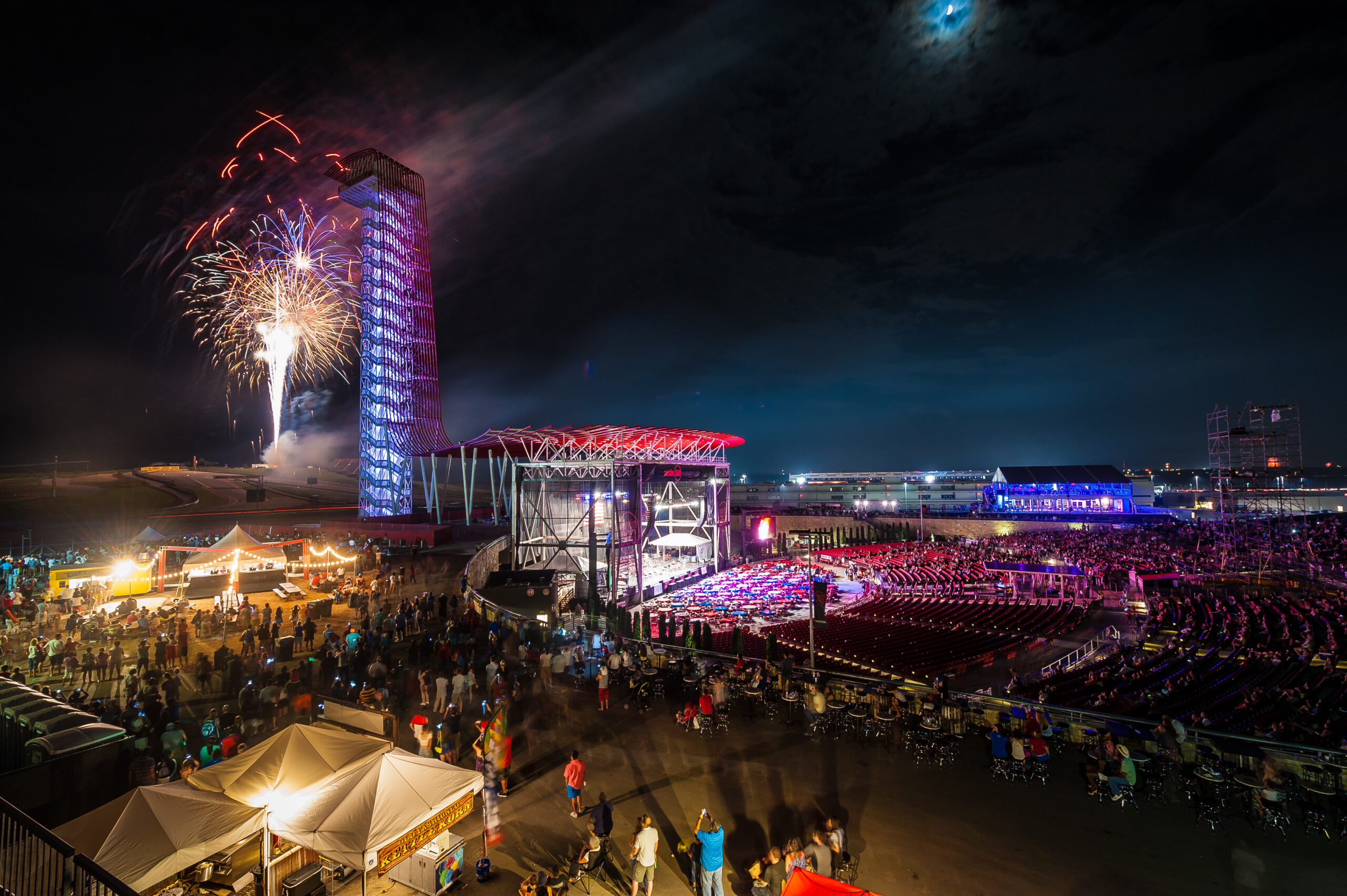 A nighttime outdoor concert with a large audience, stage lighting, fireworks by a tall illuminated tower, and tents set up in the foreground under a partly cloudy sky.