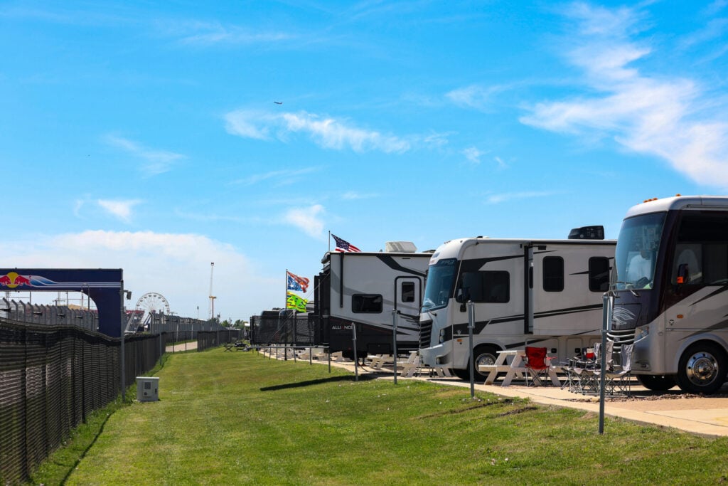 Rows of RVs are parked on a grassy area next to a racetrack; flags, chairs, and a table are set up outside under a bright, blue sky.