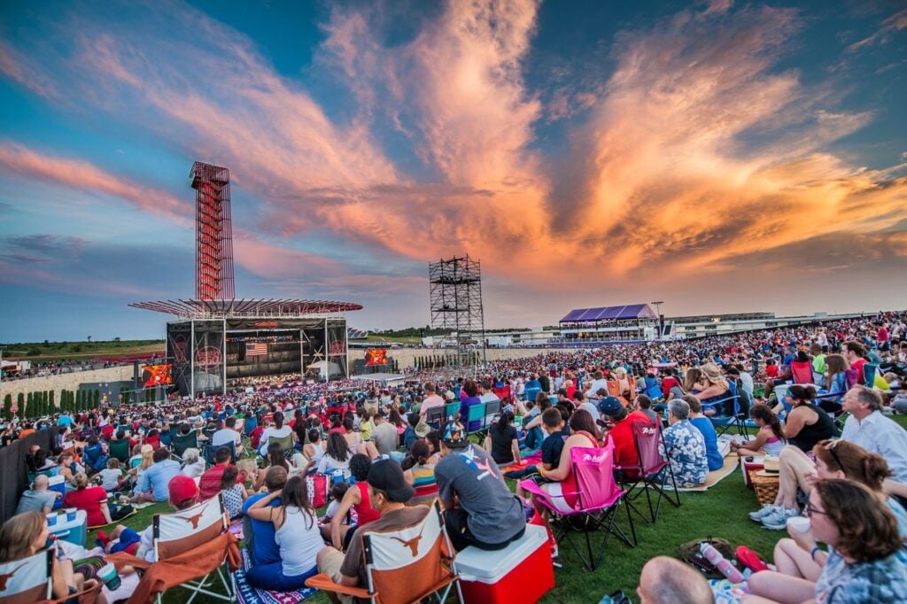 Large crowd seated on grass and in chairs faces an outdoor stage at sunset, with dramatic clouds and a red observation tower in the background.