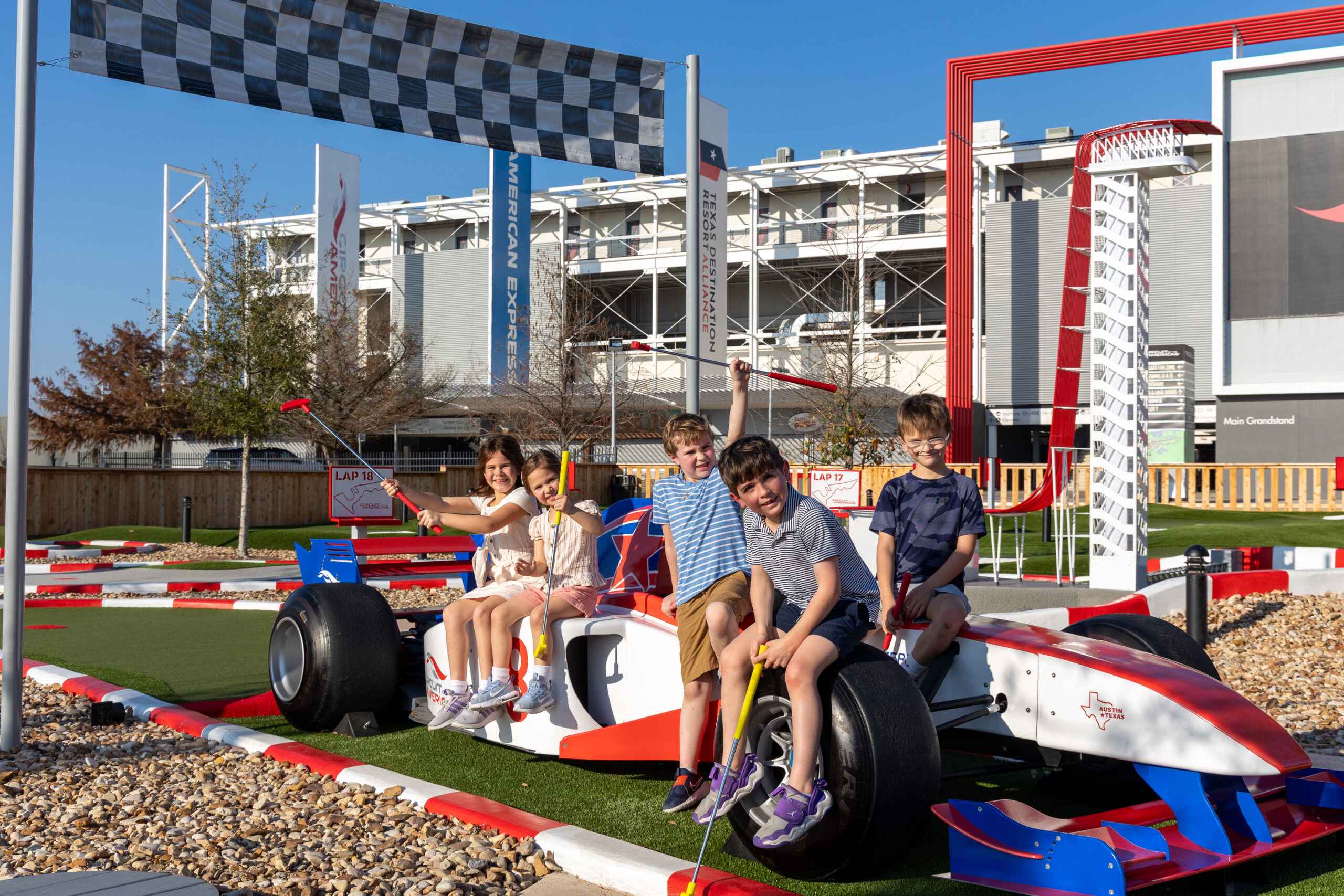 Five children sit and pose on a replica Formula 1 race car under a checkered flag at an outdoor attraction on a sunny day.