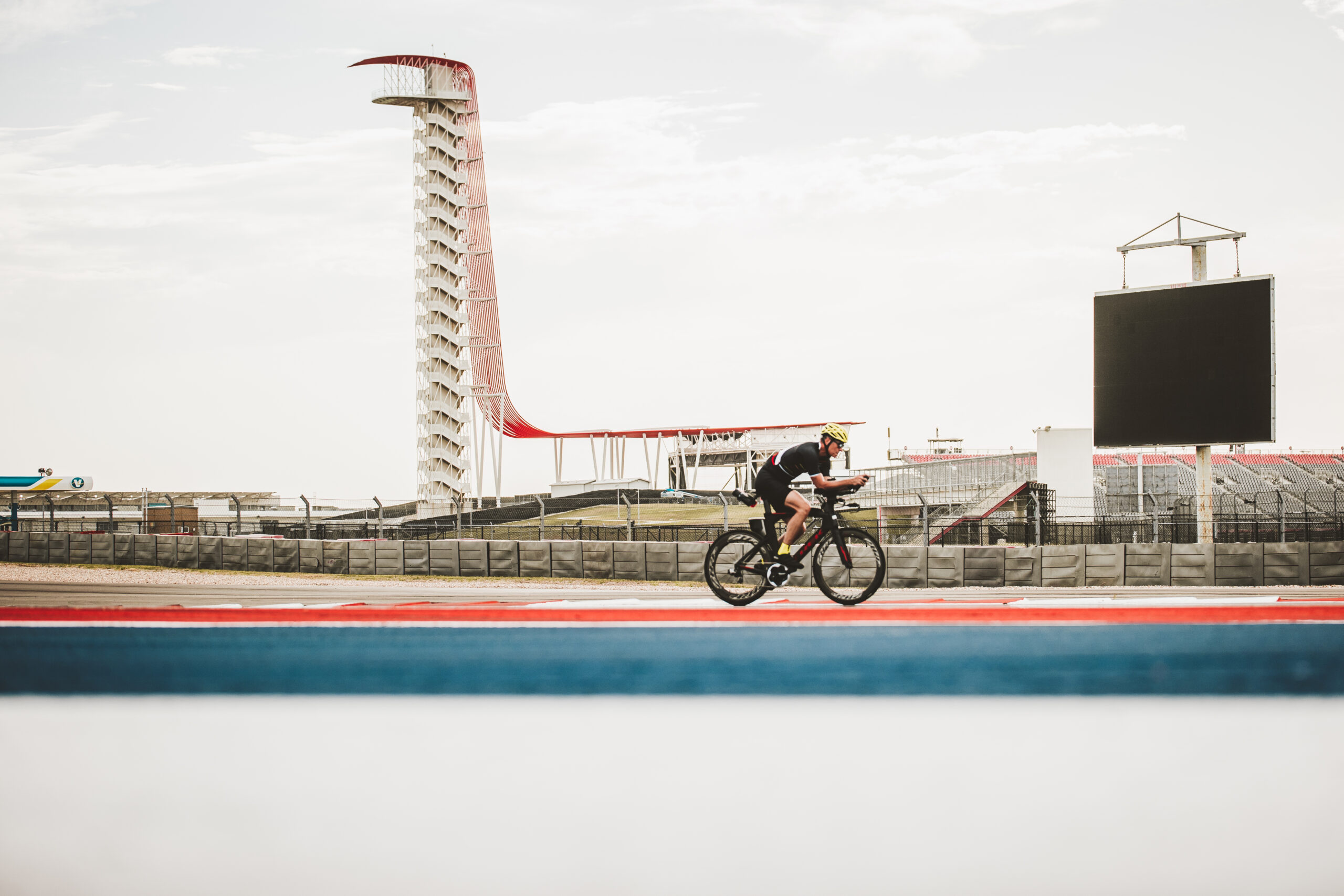 A cyclist rides on a track with a distinctive observation tower and empty grandstands in the background under a cloudy sky.