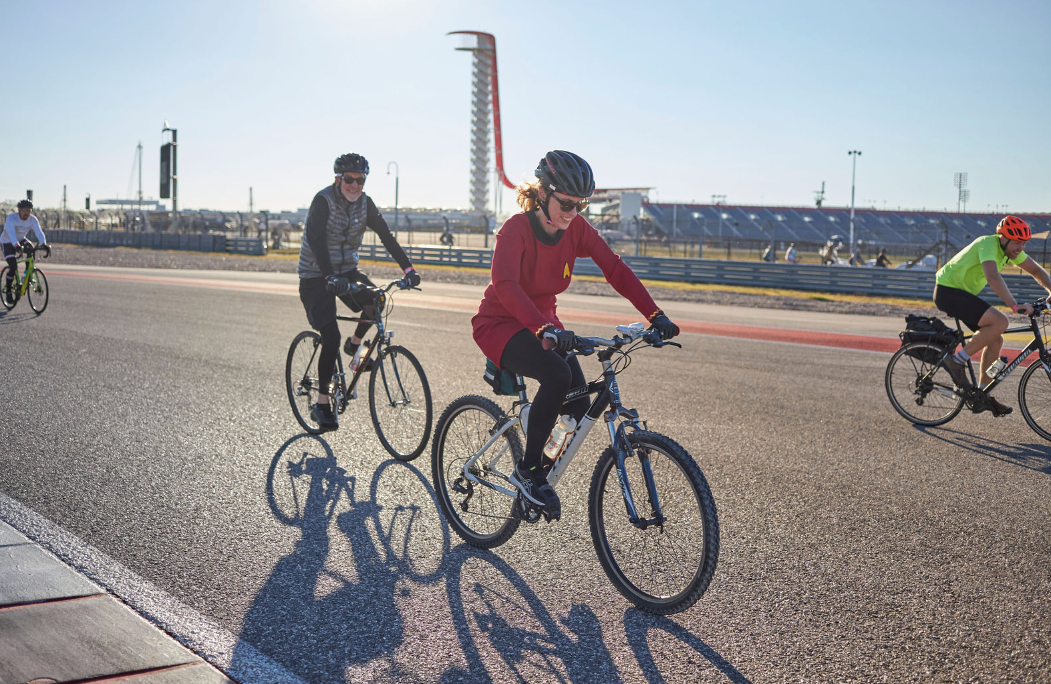 A group of people ride bicycles on a paved track under clear skies, with a tall observation tower and stadium seating visible in the background.
