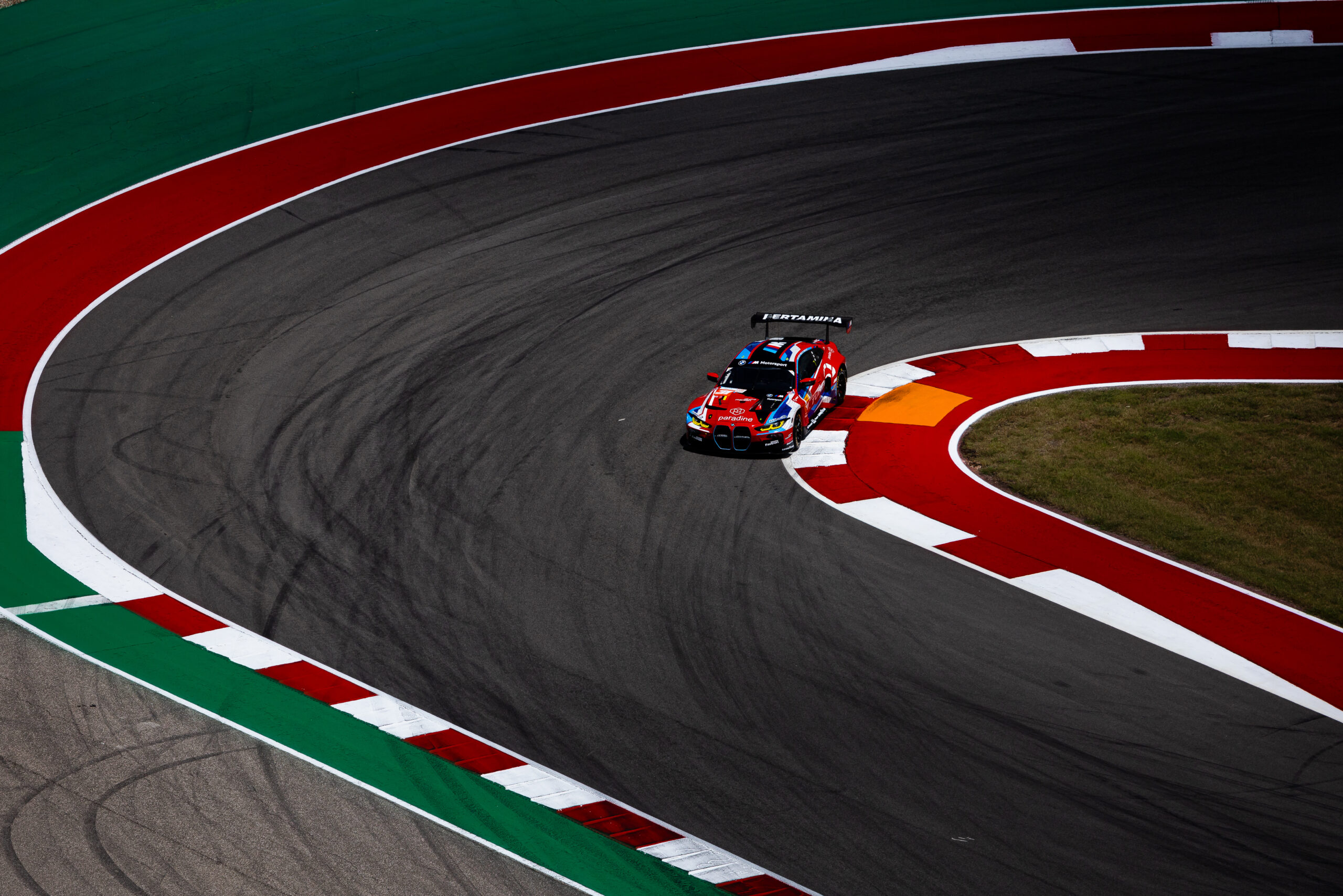 A red and blue race car drives through a sharp curve on a racetrack with red, white, and green markings.