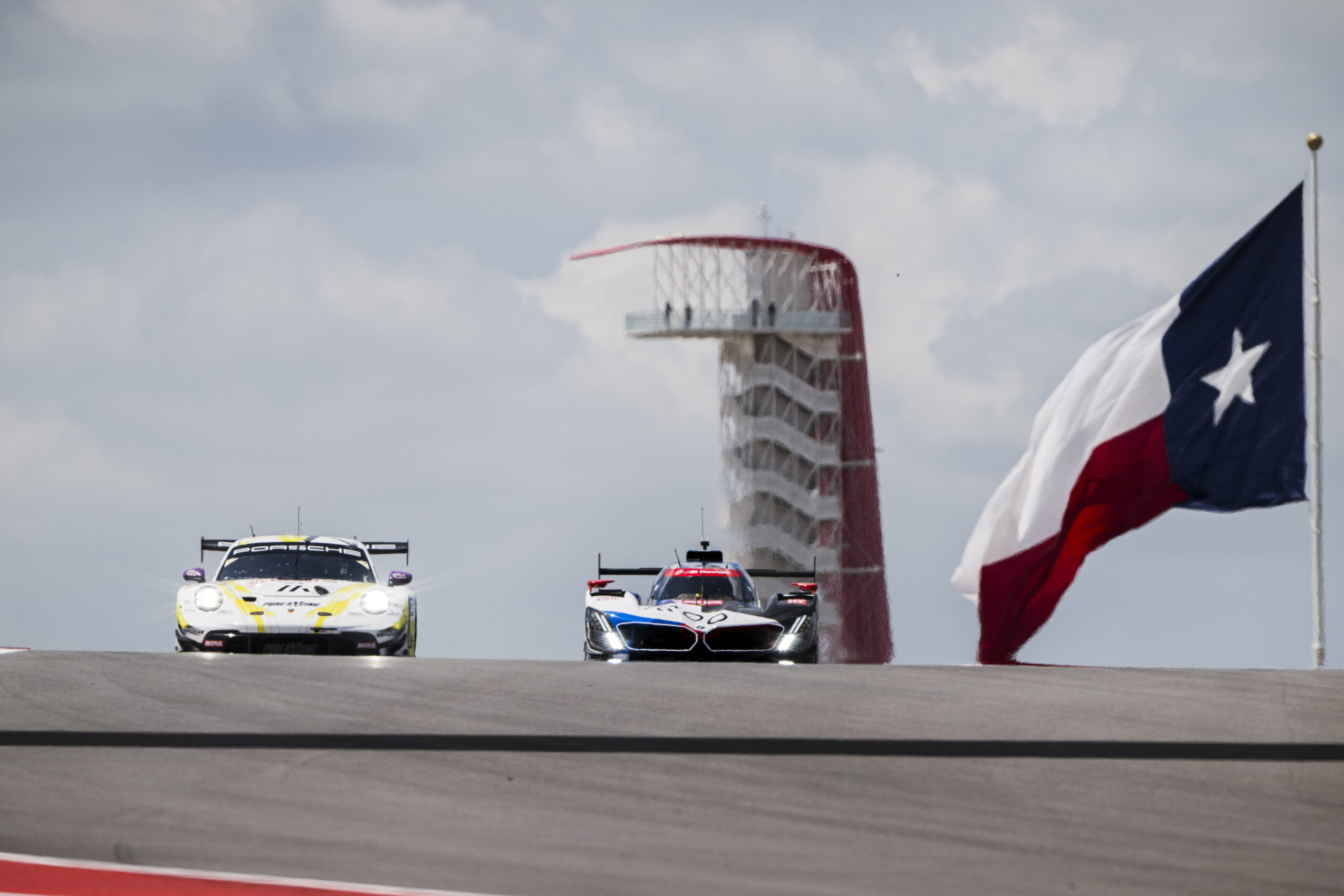 Two race cars drive side by side on a racetrack with the Texas state flag and the Circuit of the Americas observation tower in the background.