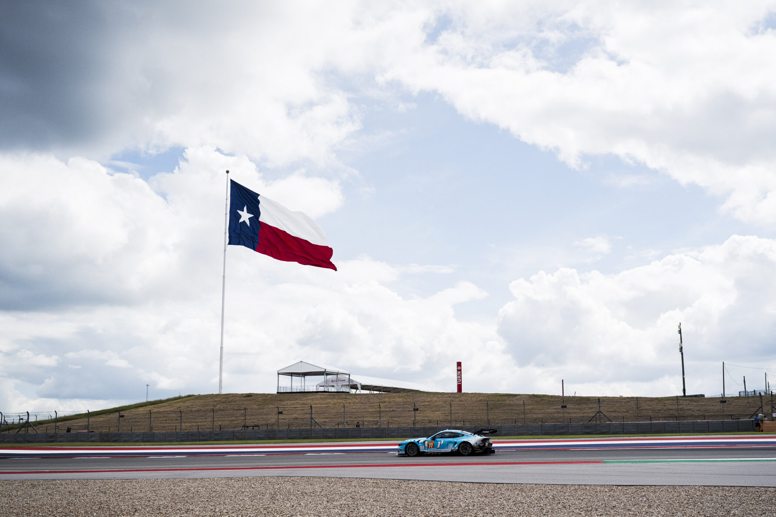 A race car drives on a track beneath a large Texas state flag on a cloudy day.