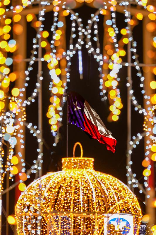 A blurred U.S. flag waves in the background, framed by bright yellow and white holiday lights and illuminated decorations.