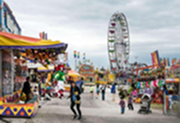 People walk through a busy outdoor carnival with game booths, colorful tents, and a large Ferris wheel in the background under a cloudy sky.