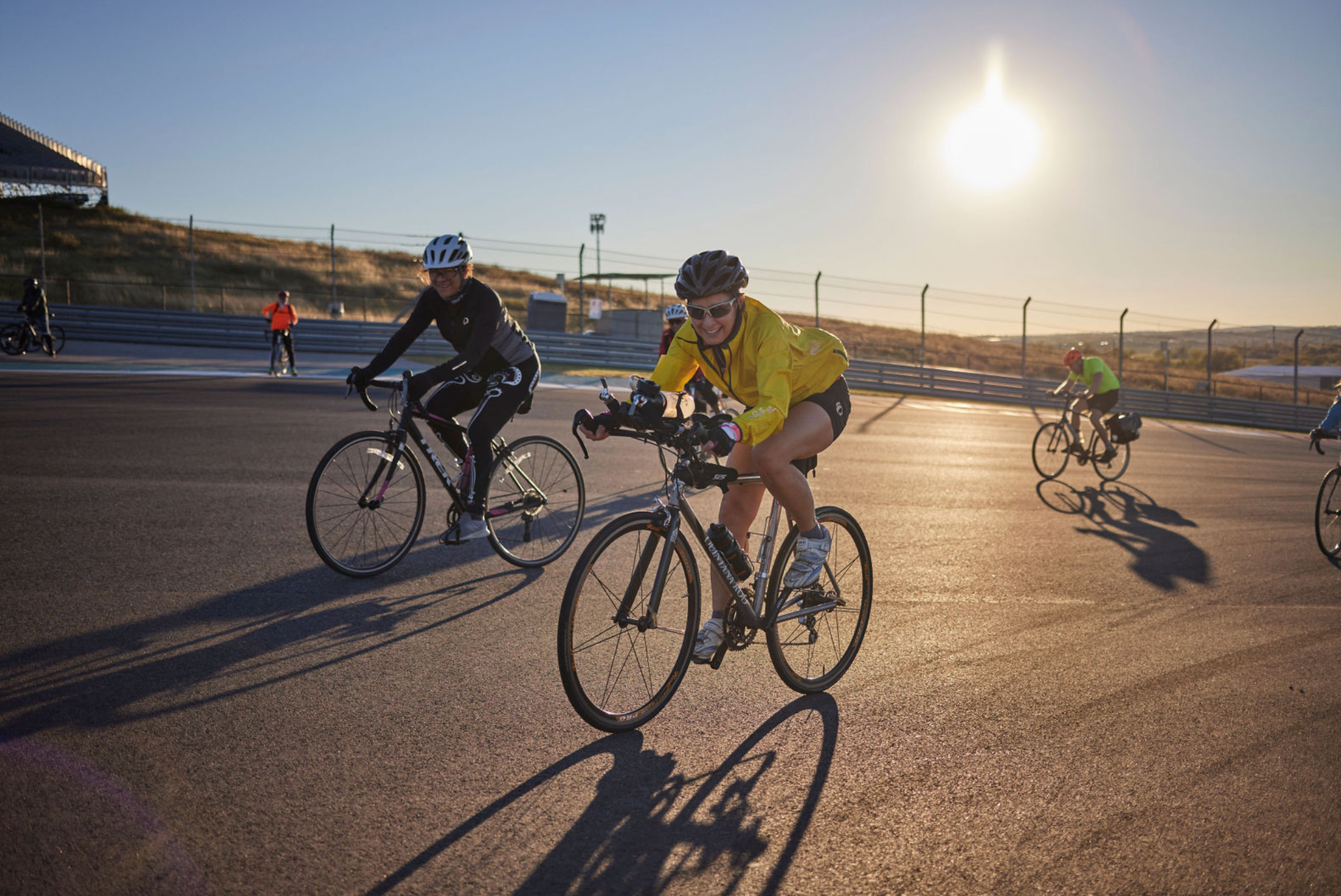 Cyclists ride on a paved track at sunrise or sunset, wearing helmets and sports gear, with a fenced area and stadium seating in the background.