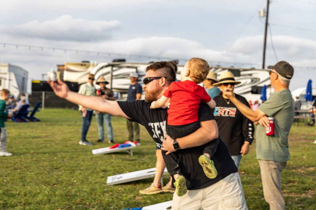 A man holding a small child throws a bean bag during an outdoor cornhole game. Several people are standing and watching, with RVs and chairs in the background.