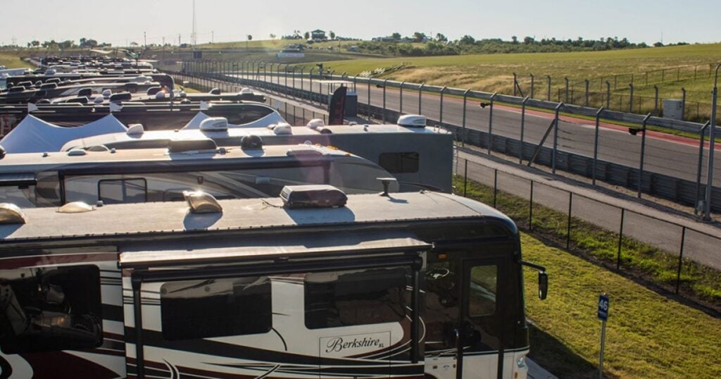 Rows of RVs are parked beside a fenced racetrack on a sunny day, with grassy hills and a clear sky in the background.