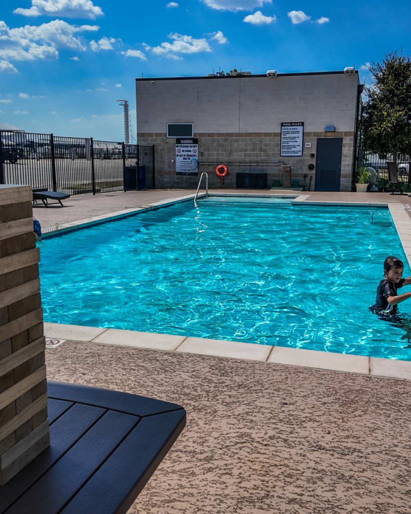 Outdoor swimming pool with clear water, surrounded by a fence, a building with pool rules, and one person standing in the water under a sunny sky.